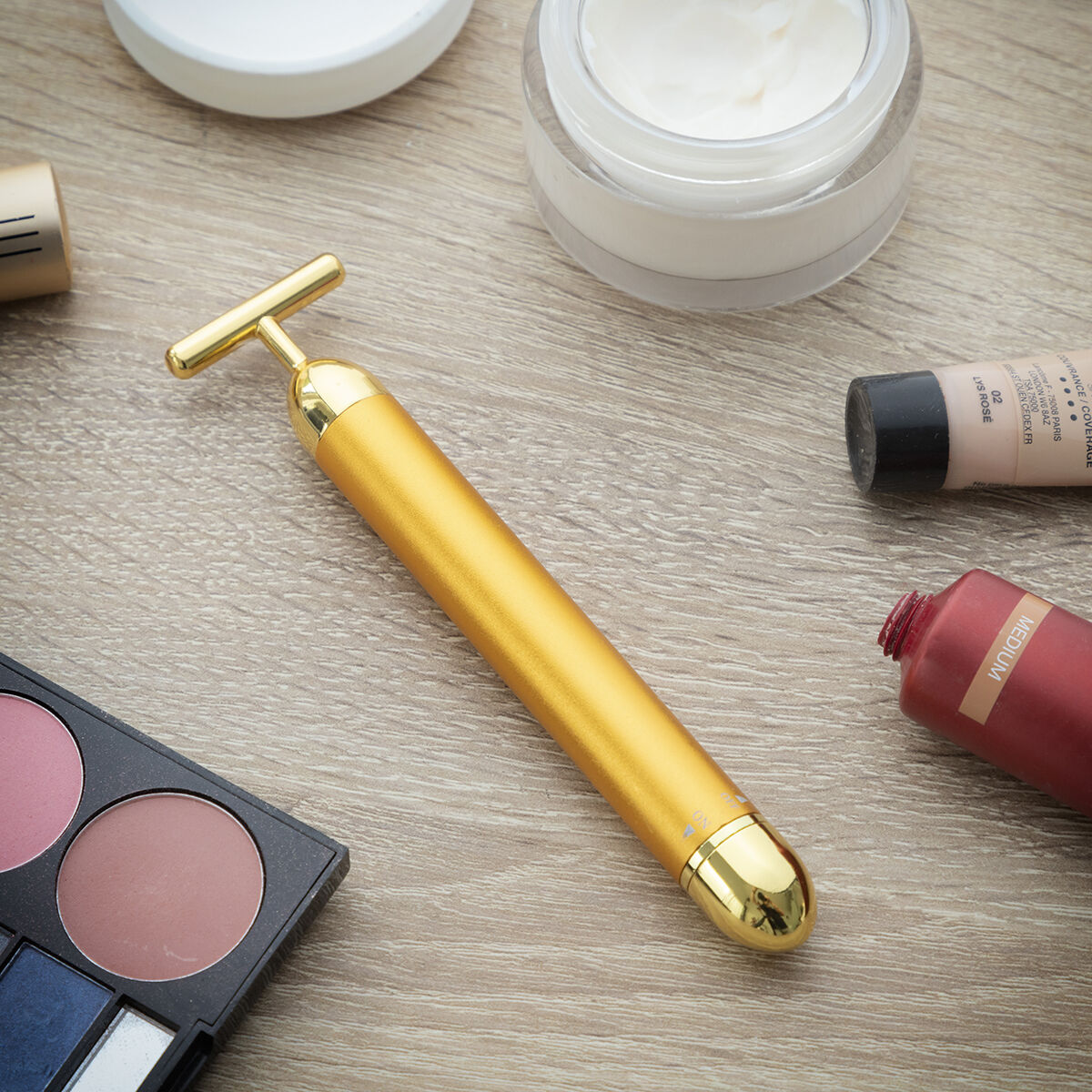 Un massaggiatore facciale dorato è appoggiato su un tavolo con prodotti per il trucco. Il massaggiatore ha una forma a T.