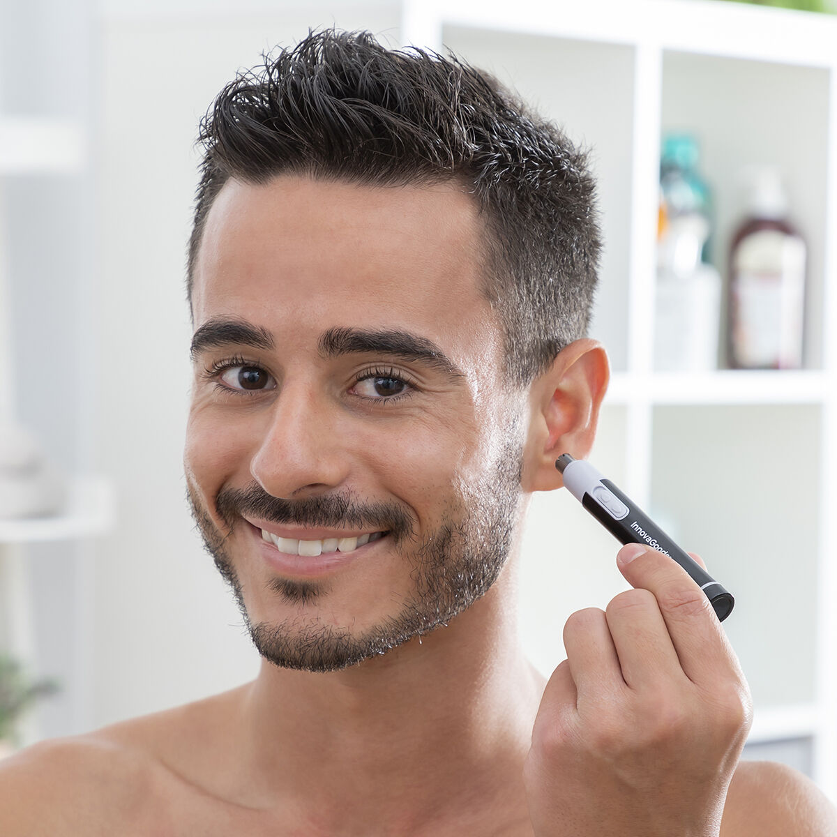 Un hombre se está recortando el vello de la oreja con un pequeño recortador blanco y negro. Está sonriendo.