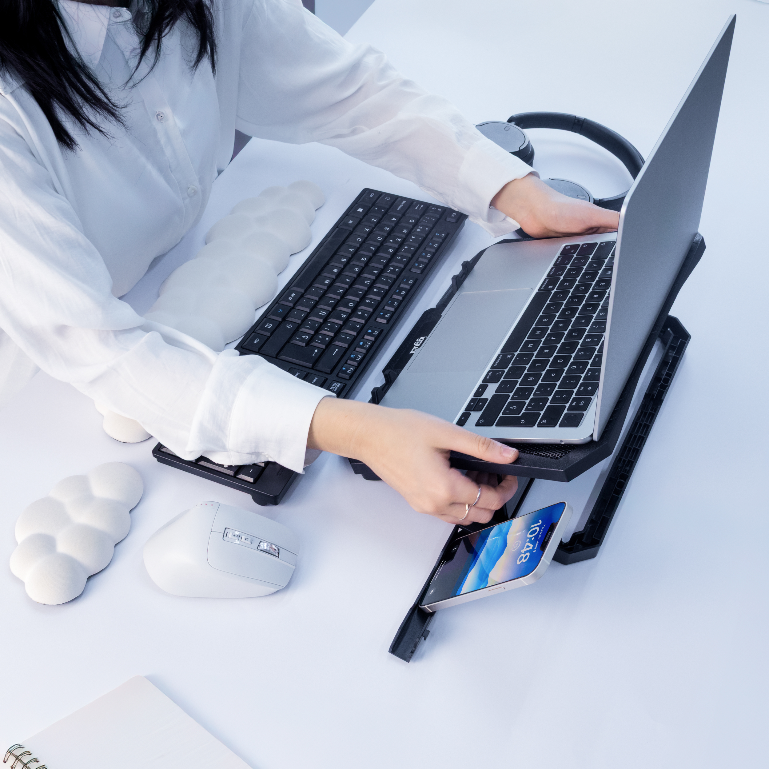 Osoba korzystająca z laptopa na stojaku chłodzącym, z klawiaturą, myszą i telefonem na białym biurku.