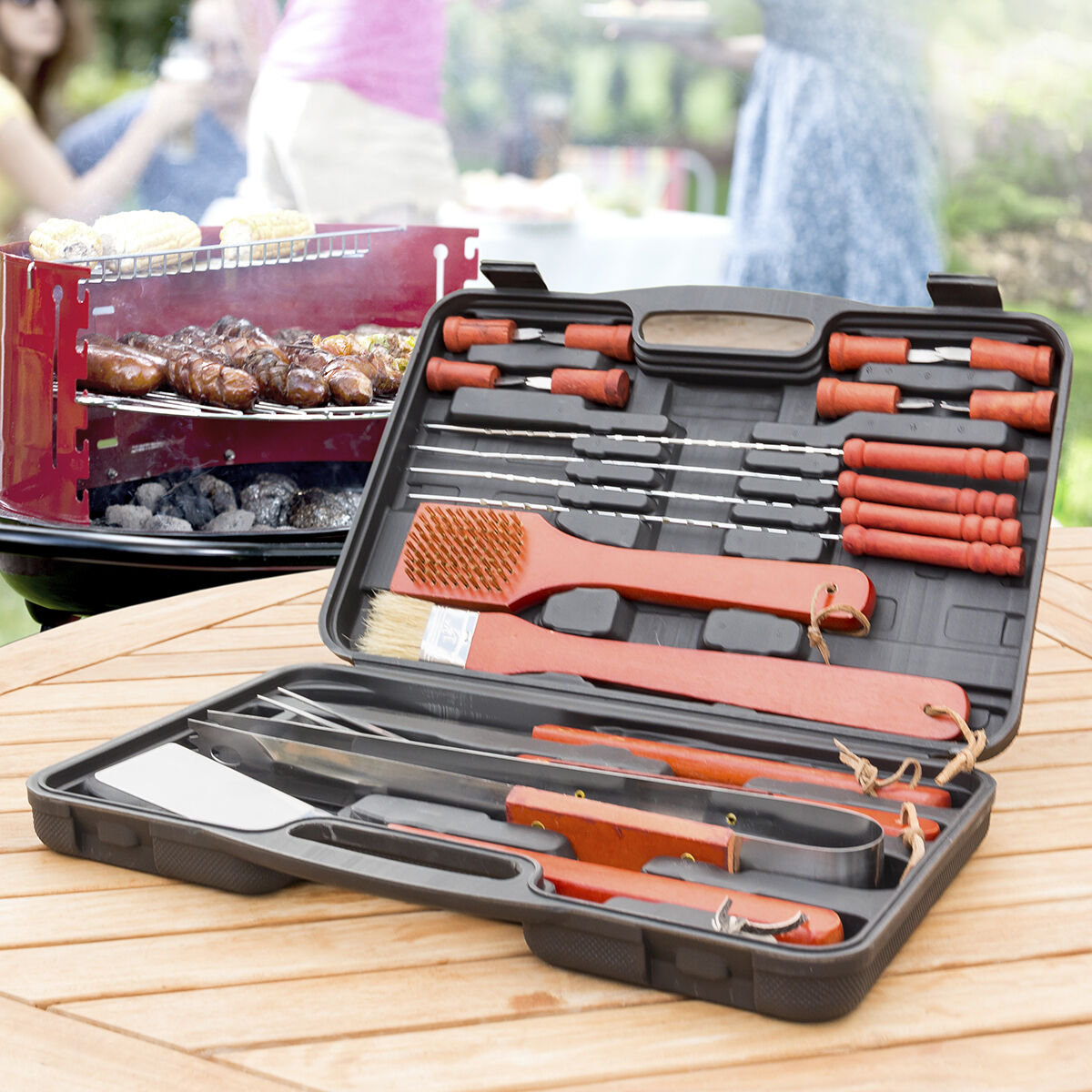 Eine schwarze Werkzeugkiste mit roten Grillwerkzeugen ist auf einem Holztisch geöffnet. Personen und ein Grill sind im Hintergrund.