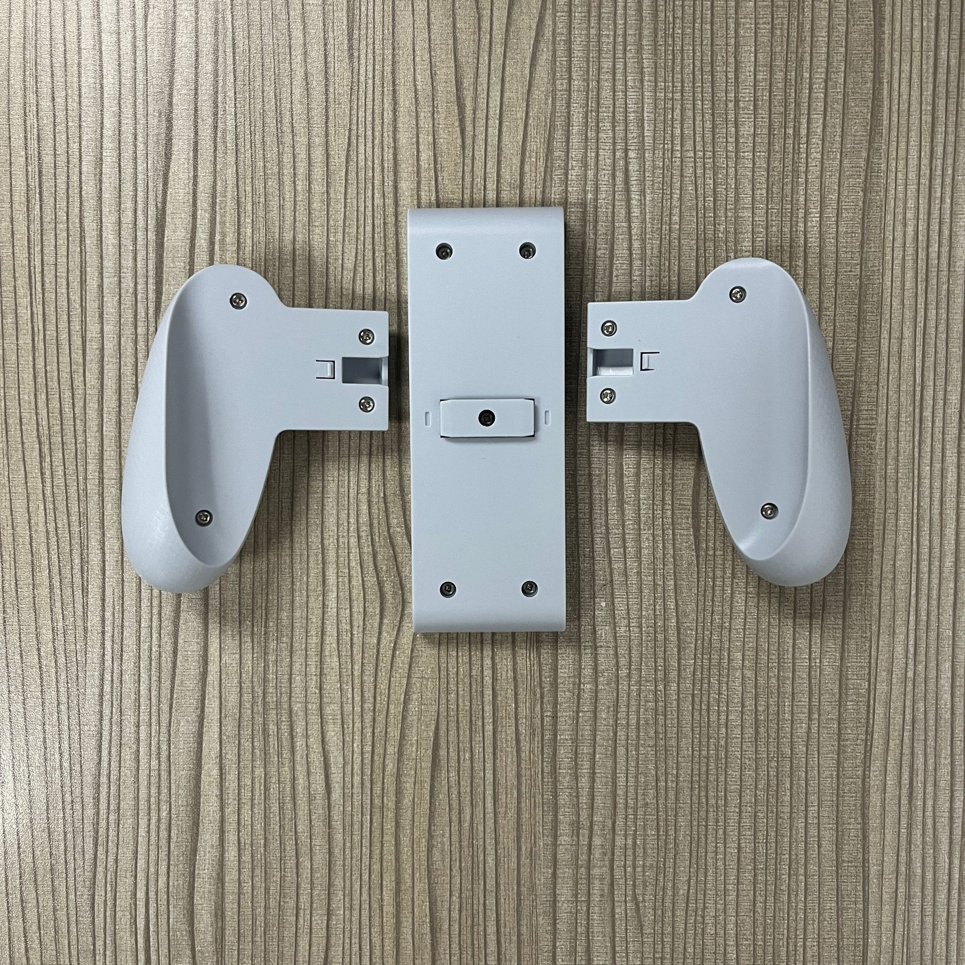 Three detached white game controller parts on a wood-textured surface. Center piece has screws, side pieces are curved.