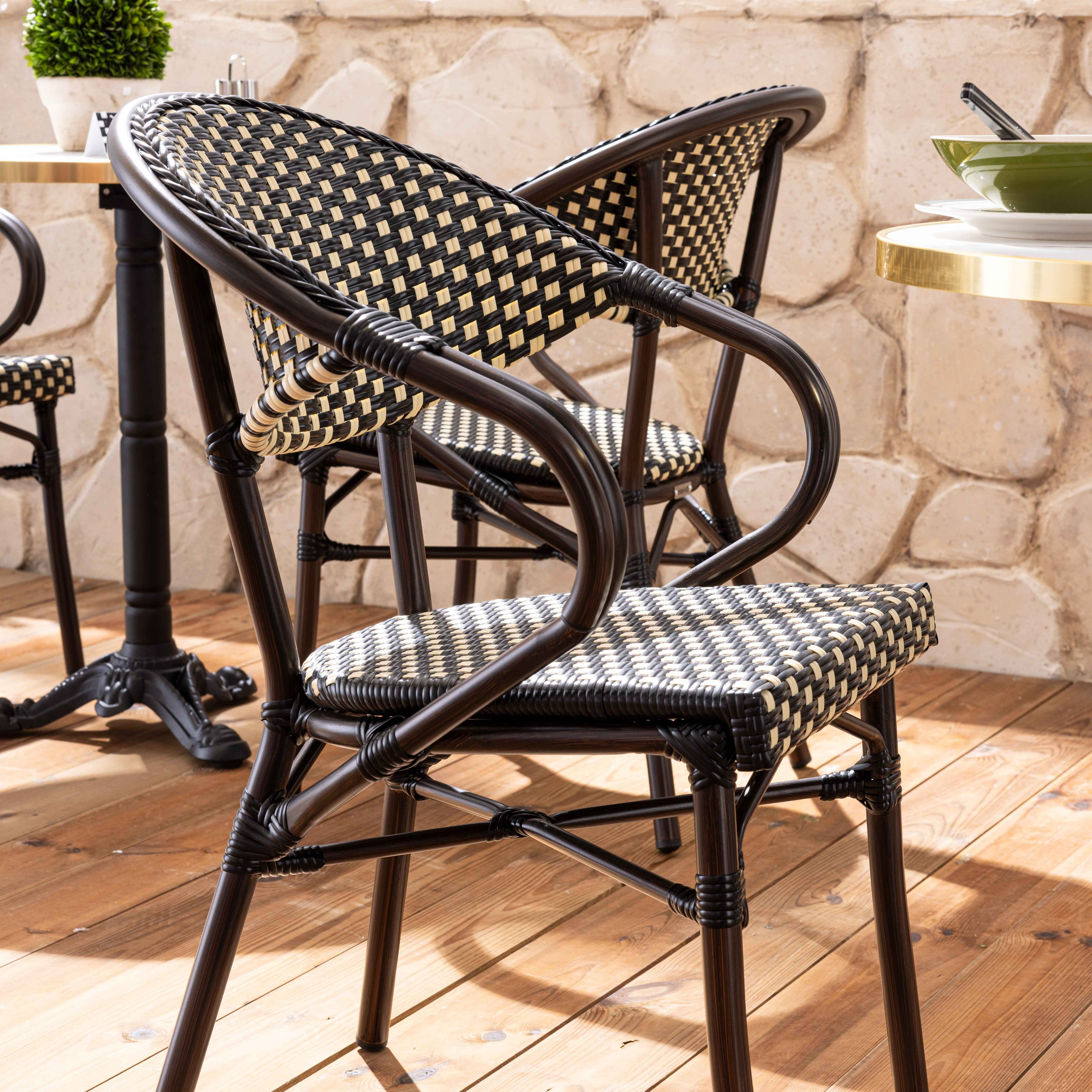 Zwei Rattanstühle mit schwarz-beige gewebten Sitzen und Rückenlehnen stehen auf einer Holzterrasse in der Nähe von Steinmauern.