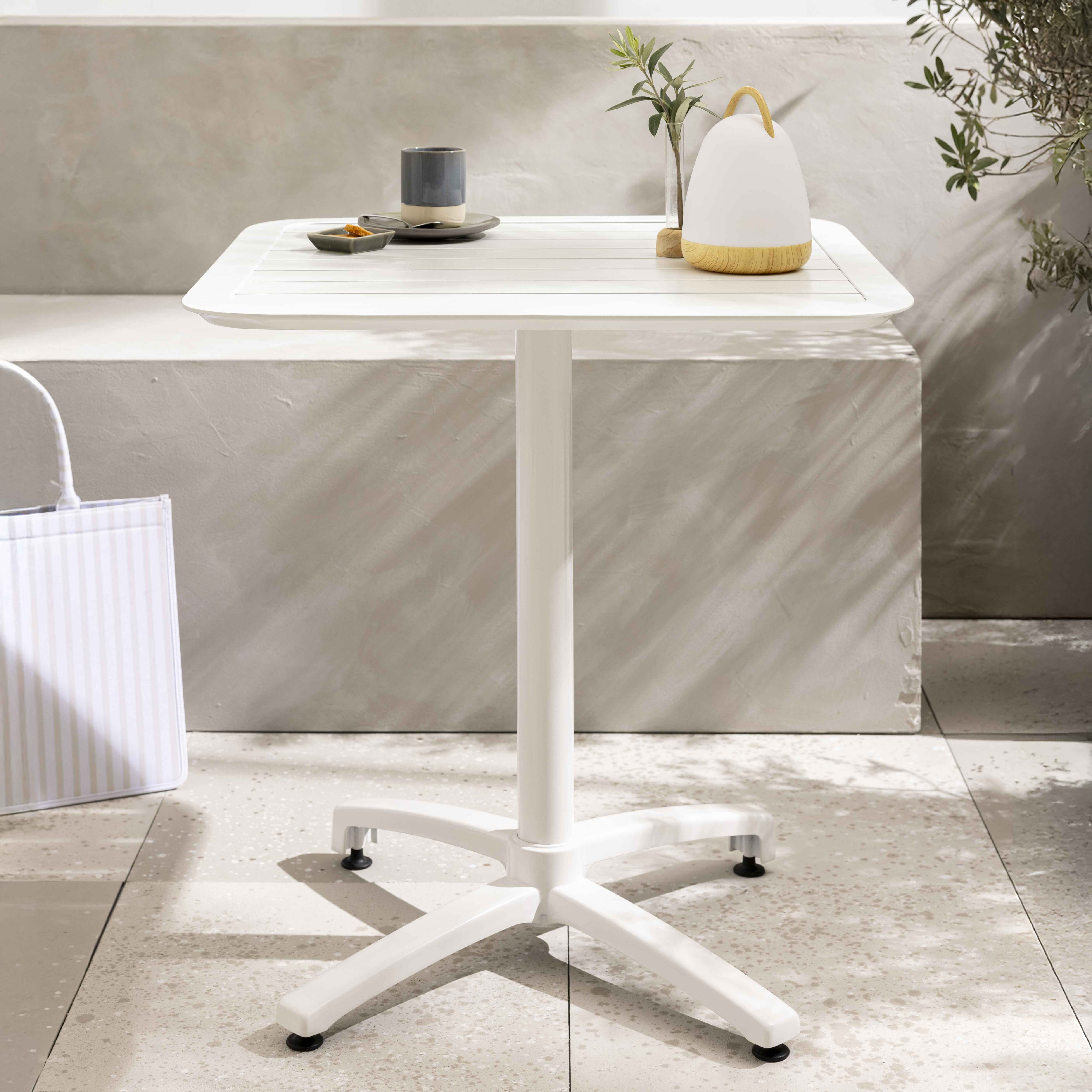 Ein weißer Outdoor-Tisch mit Lampe, Trinkgeschirr und einer Tragetasche auf einer Terrasse, vor einer strukturierten Wand und Blattwerk.