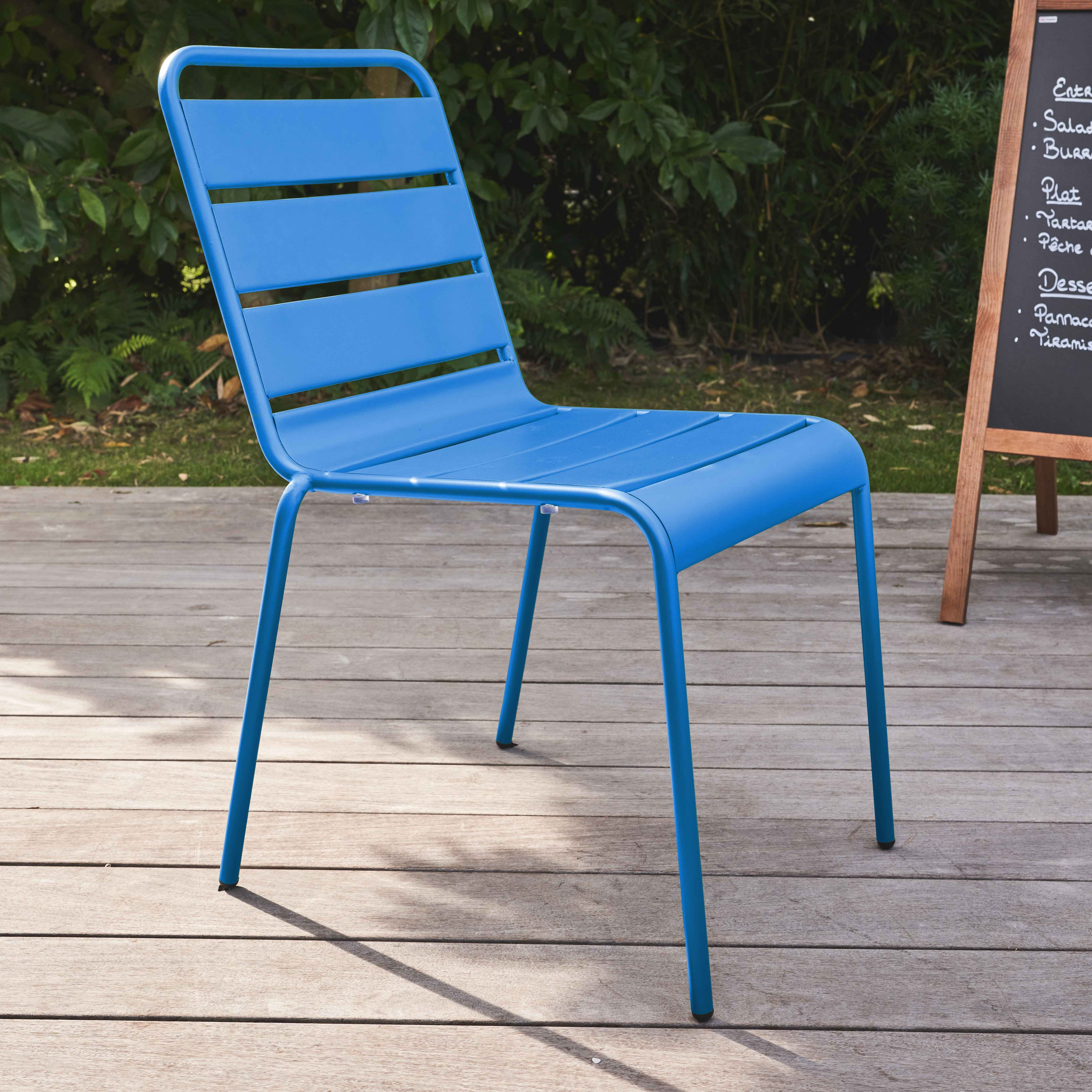 Ein blauer Stuhl mit einer Lattenrostlehne und vier Beinen steht auf einer Holzterrasse mit sichtbarem Grün.