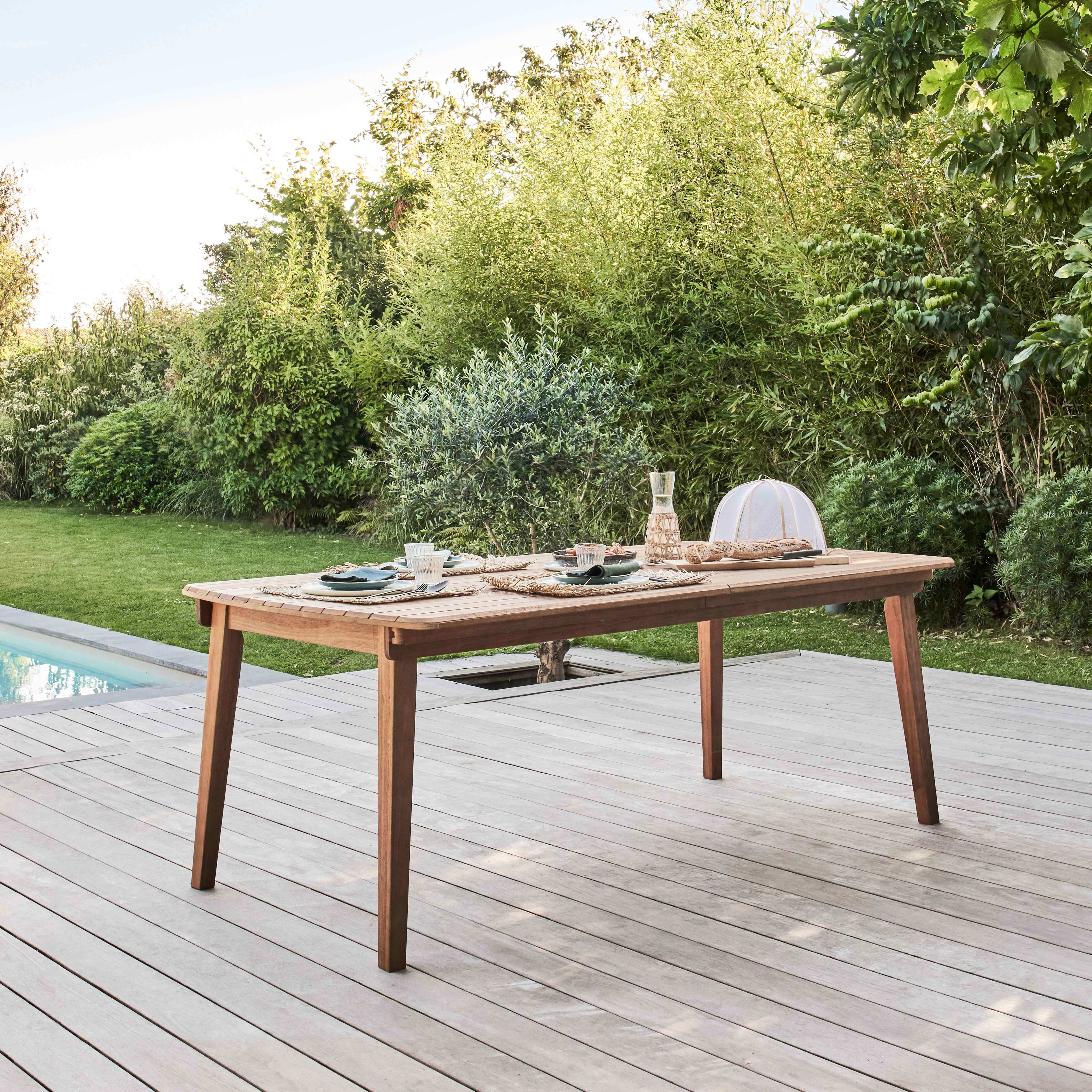 Holz-Esstisch für vier Personen im Freien auf einer Terrasse mit Pool und Bäumen im Hintergrund.