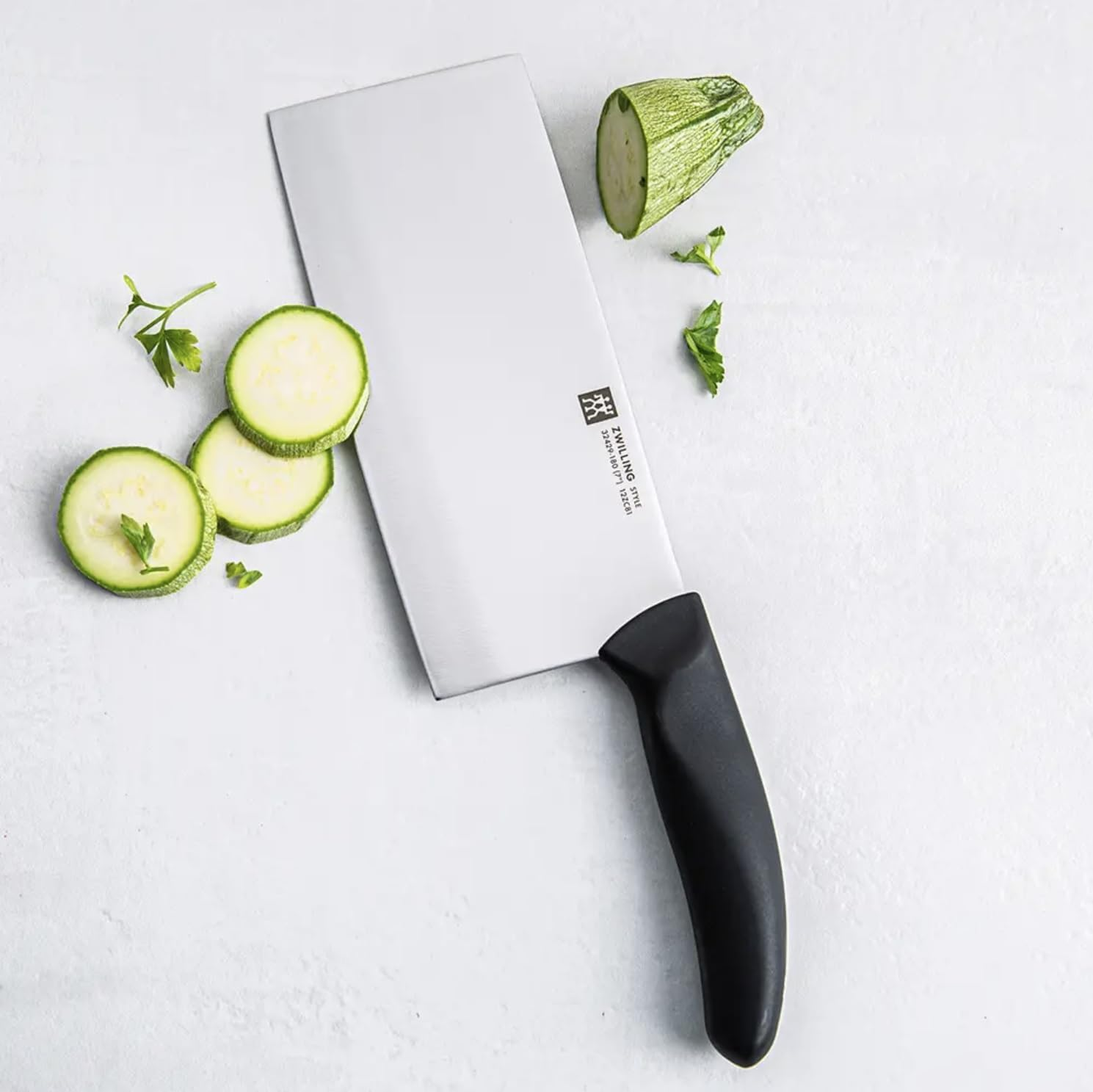 Ein schwarzes Hackmesser auf einer weißen Oberfläche mit gehackten Zucchini und Petersilie.