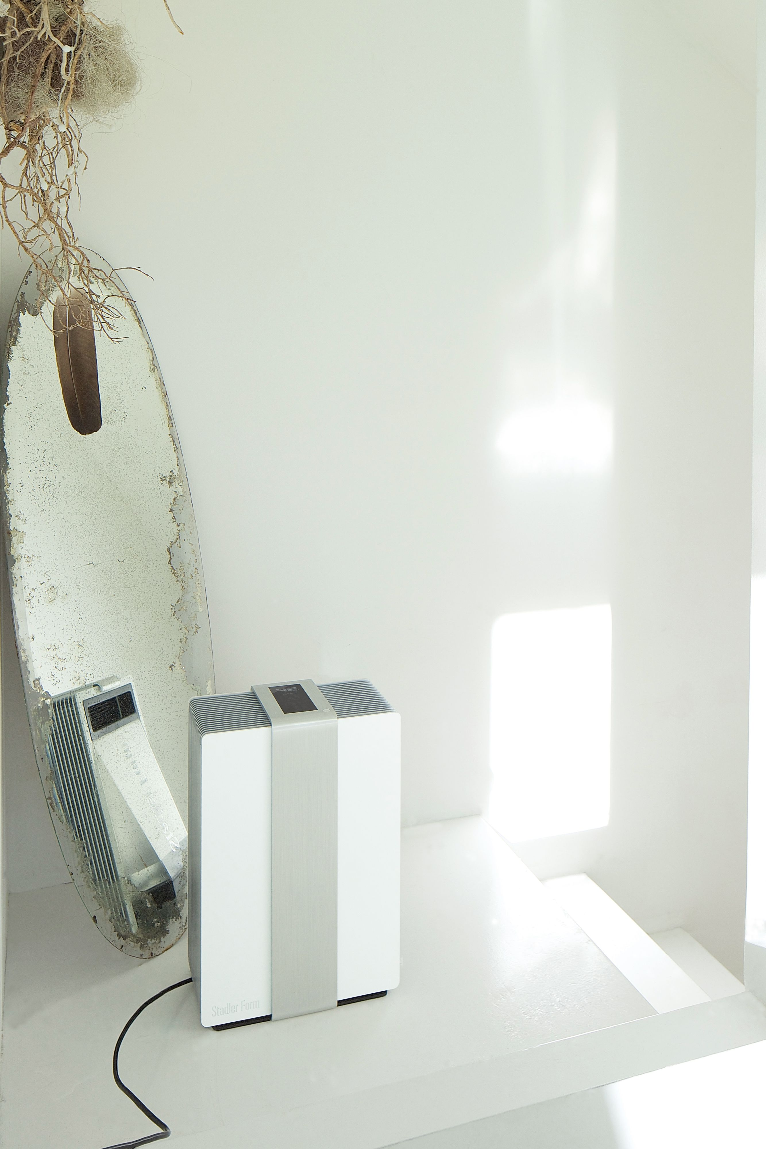 White air purifier near a mirror. Room is white, with sunlight. Black power cable.