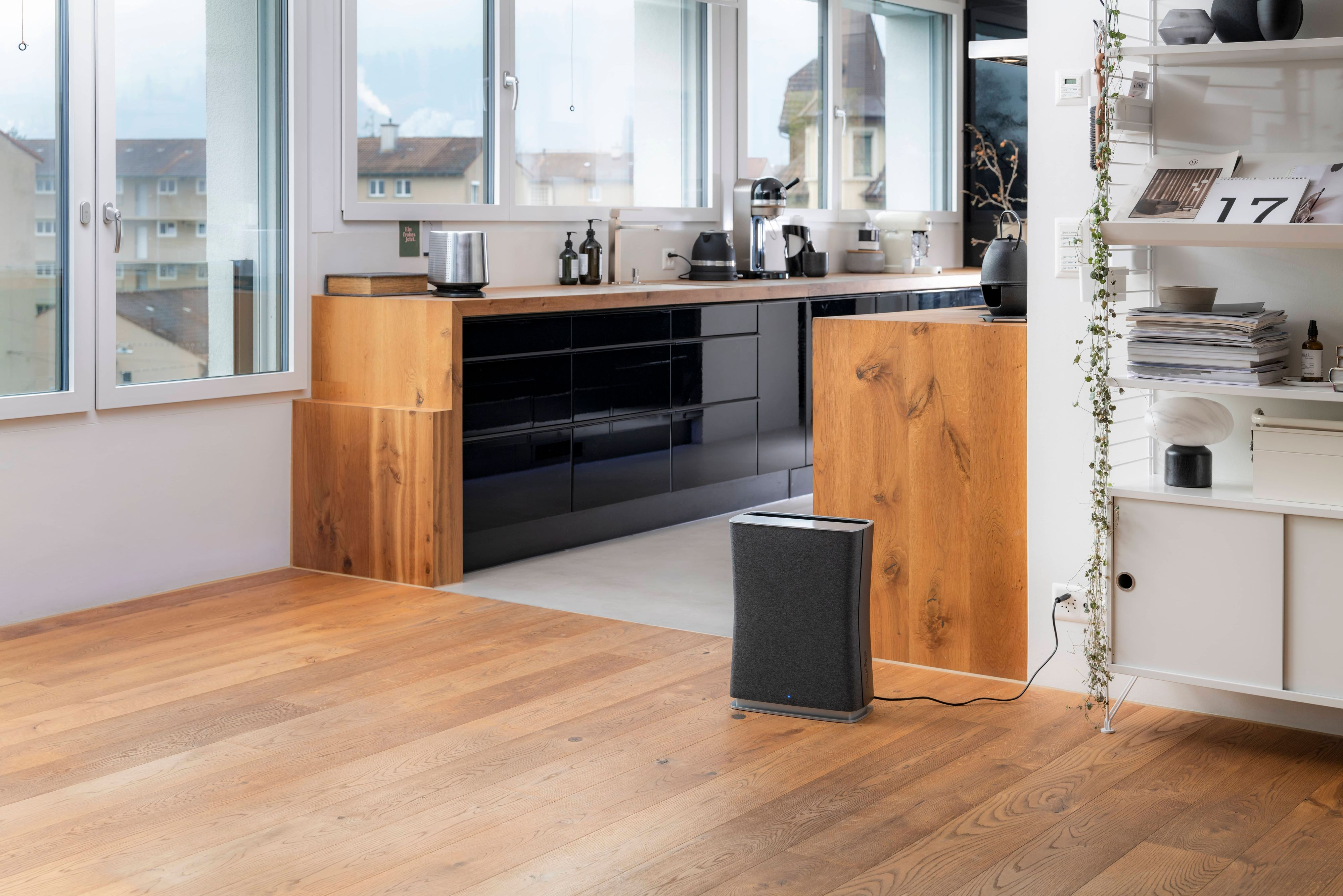 Kuchnia z drewnianą podłogą, czarnym blatem, oknami. Ciemnoszary oczyszczacz powietrza stoi na podłodze.