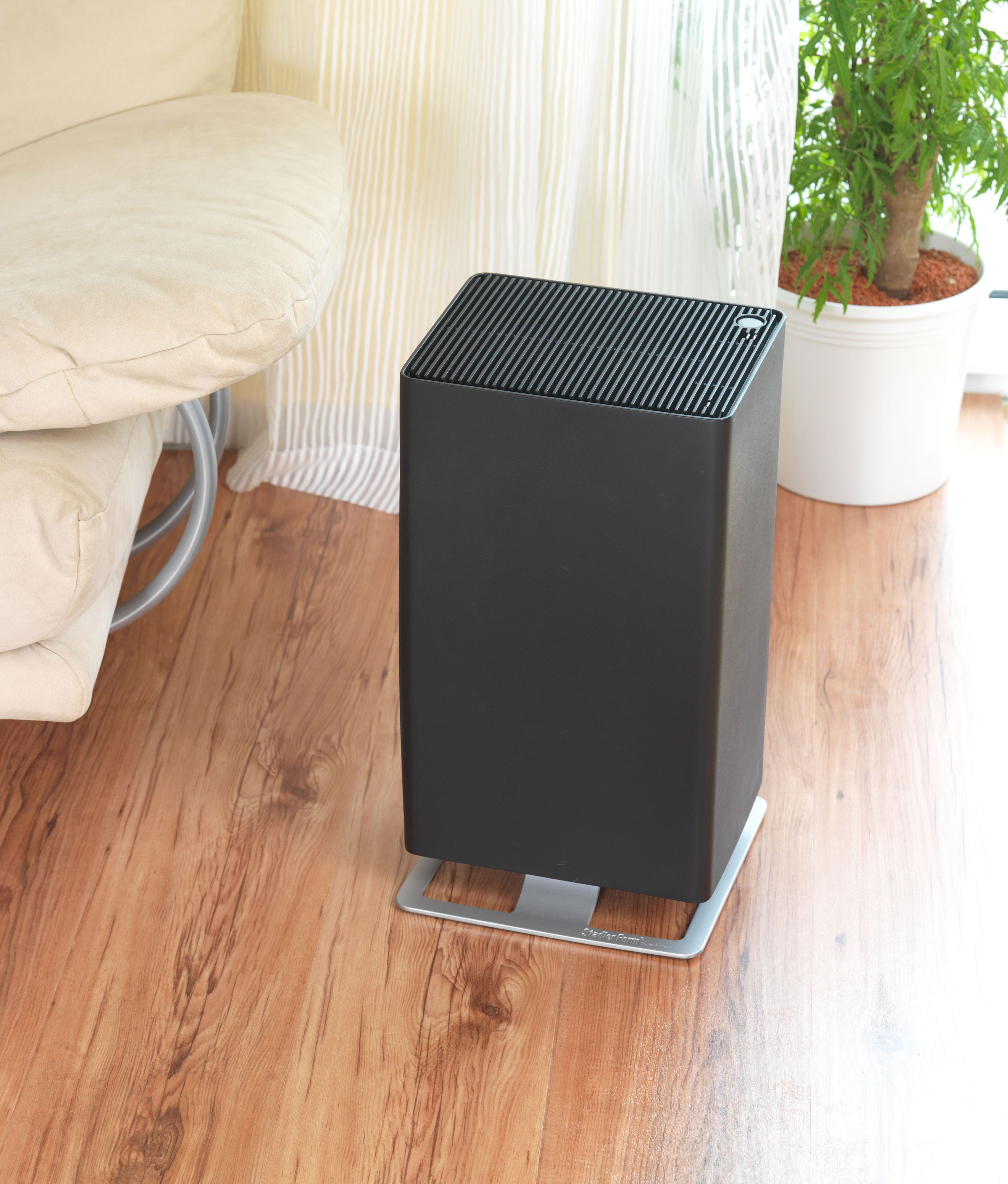 Purificatore d'aria nero su pavimento in legno, con divano beige e pianta sullo sfondo.