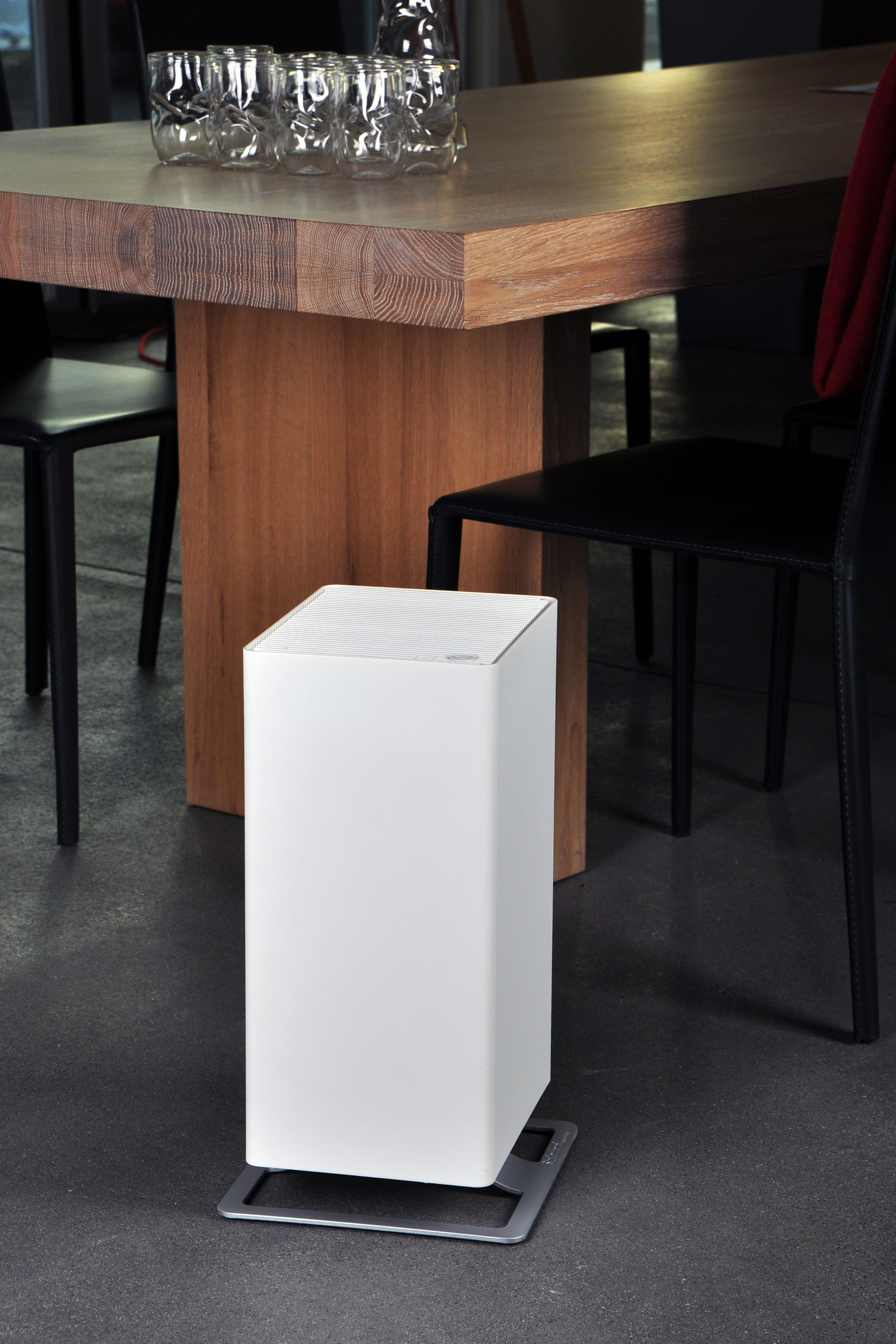 A white air purifier stands near a table, chairs, and a grey floor.