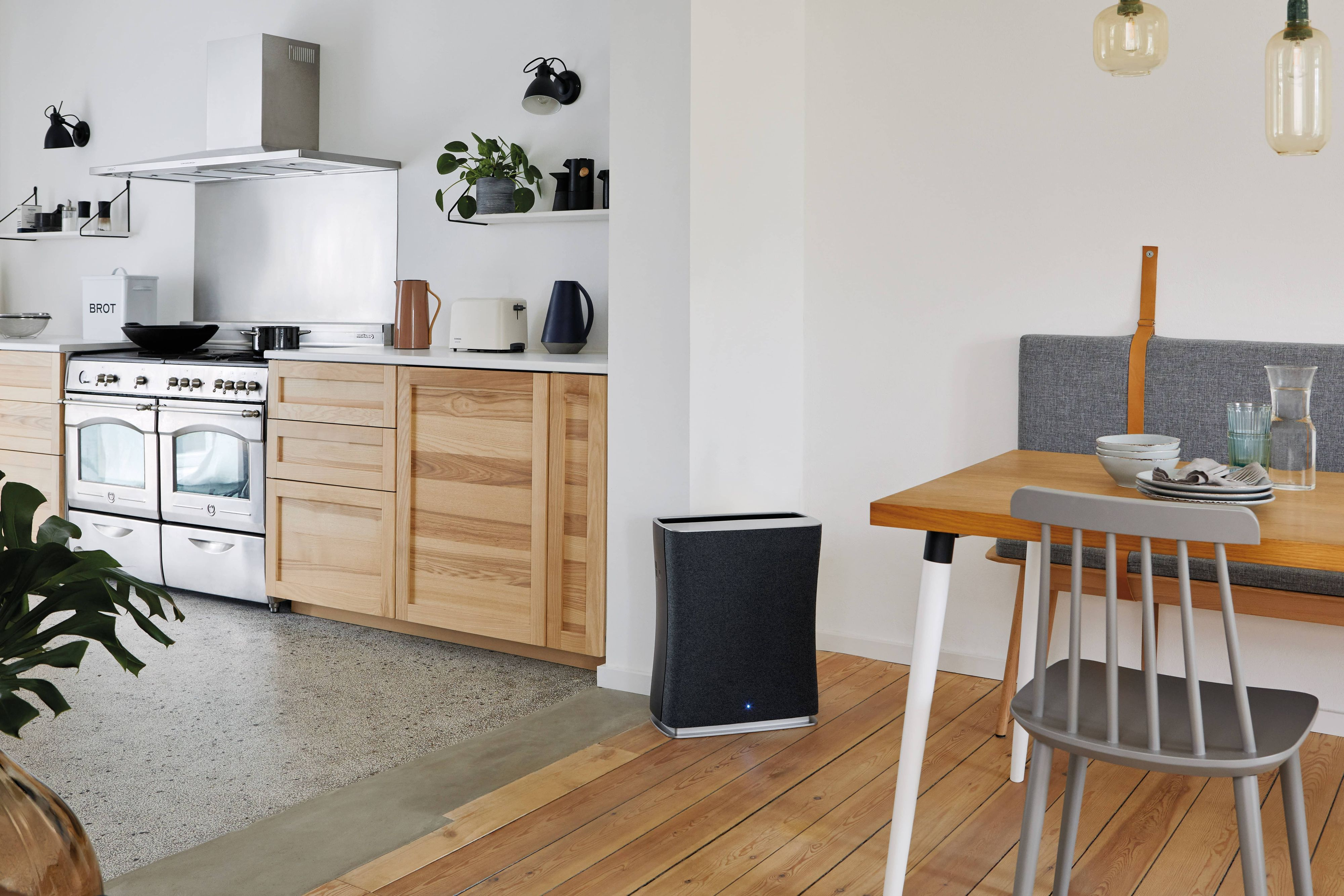 An air purifier stands in a kitchen with a wooden floor, near a table and cabinets.