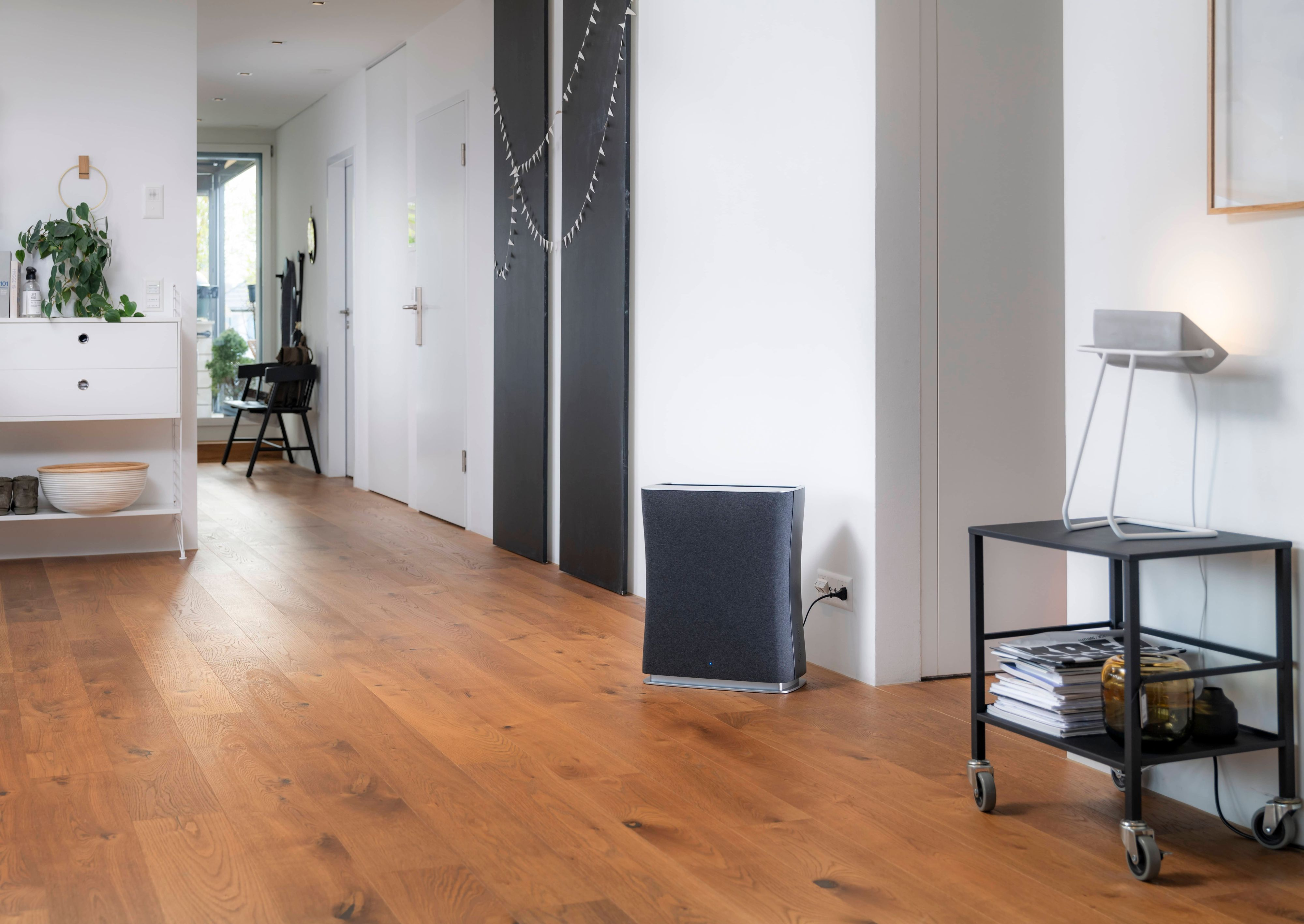 An air purifier stands in a hallway with a wooden floor, near a cart and a white door.