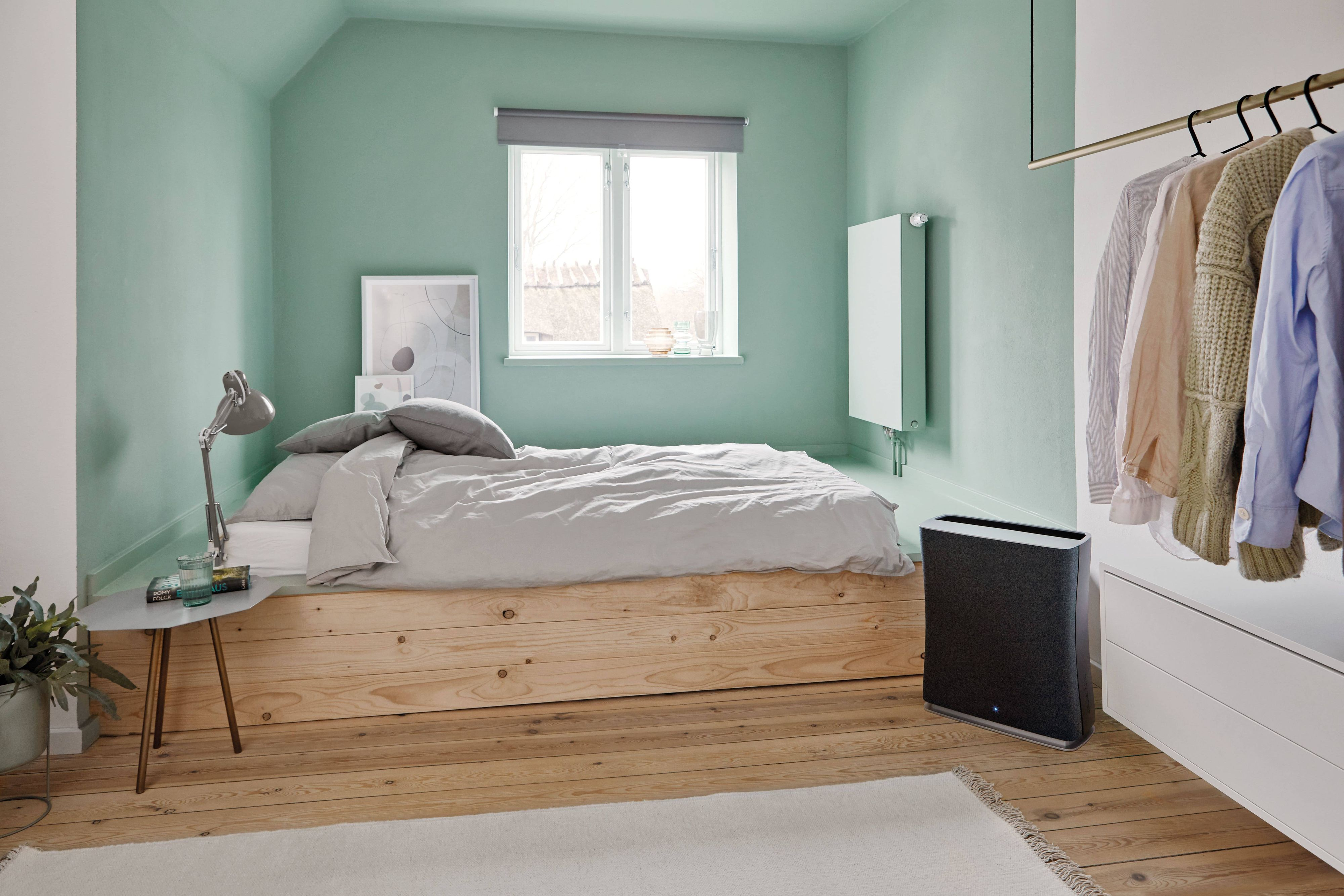 Bedroom with bed, window, clothes, and air purifier. Green walls, wooden floor, and a rug.