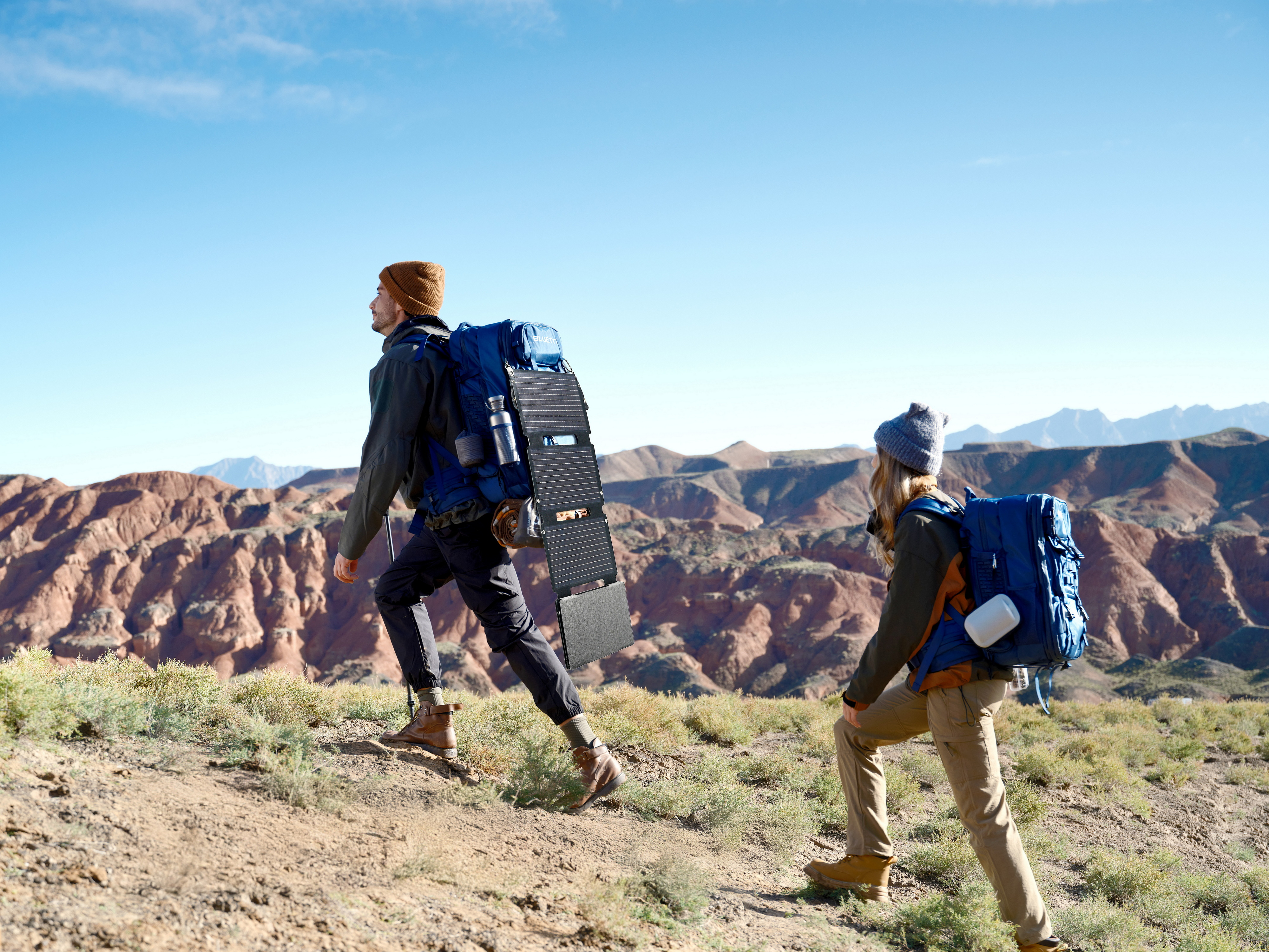 Dwóch turystów wchodzi na wzgórze z plecakami. W tle góry i błękitne niebo. Jeden ma panel słoneczny.
