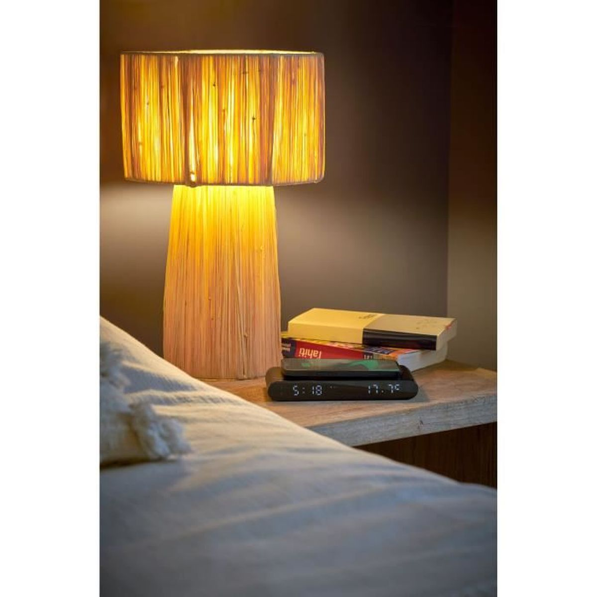 Lampa z plecionym abażurem na szafce nocnej. Łóżko i książki w tle. Obecny jest również zegar cyfrowy.