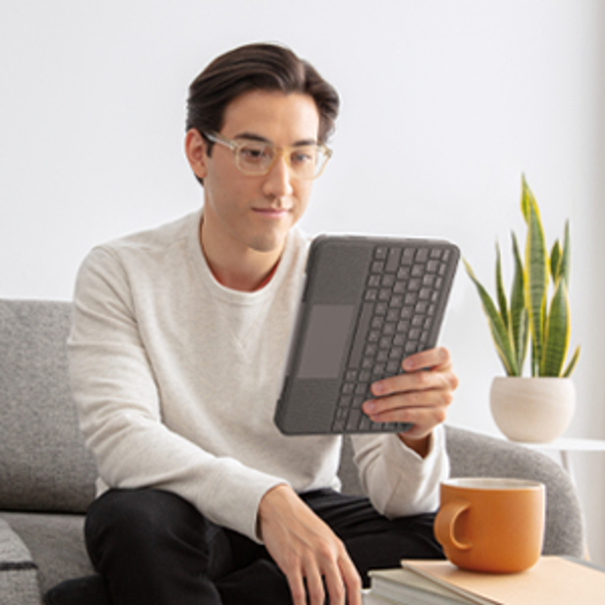Un uomo è seduto su un divano grigio, che tiene in mano un tablet con custodia per tastiera. Una tazza di caffè è su un tavolo.