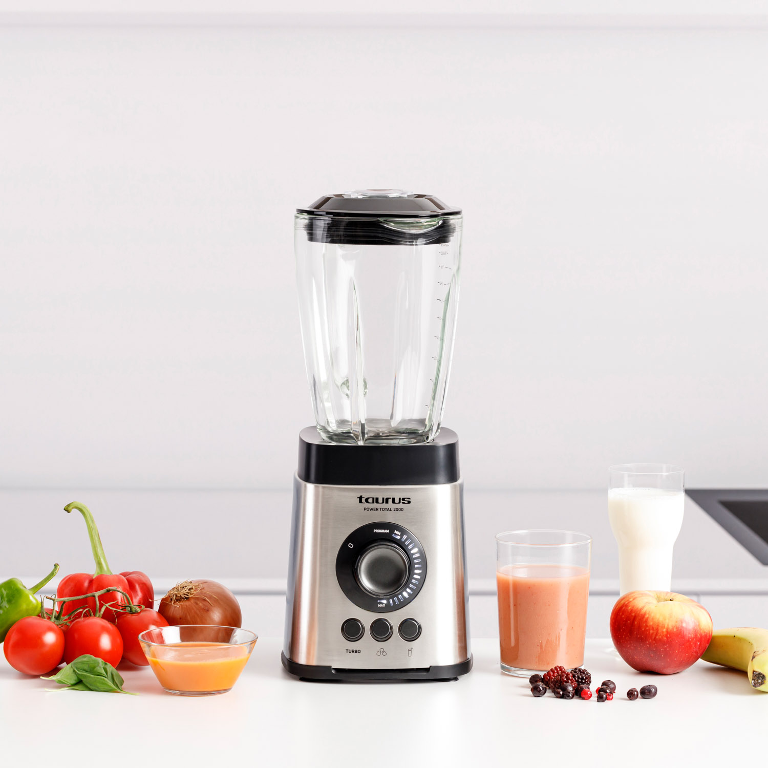 Licuadora con frutas y verduras en superficie blanca, vasos de zumo y leche.