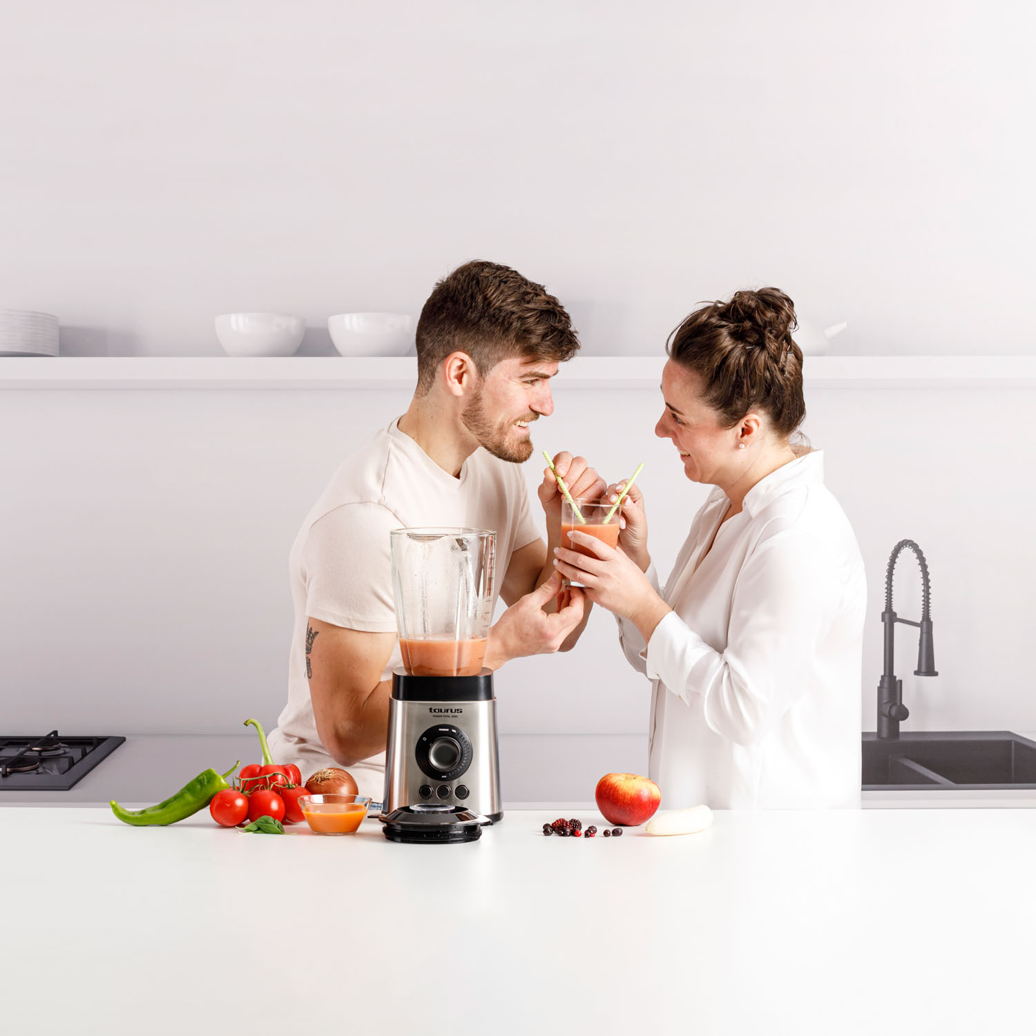 Una pareja compartiendo batido de una licuadora, con frutas y verduras en la encimera.