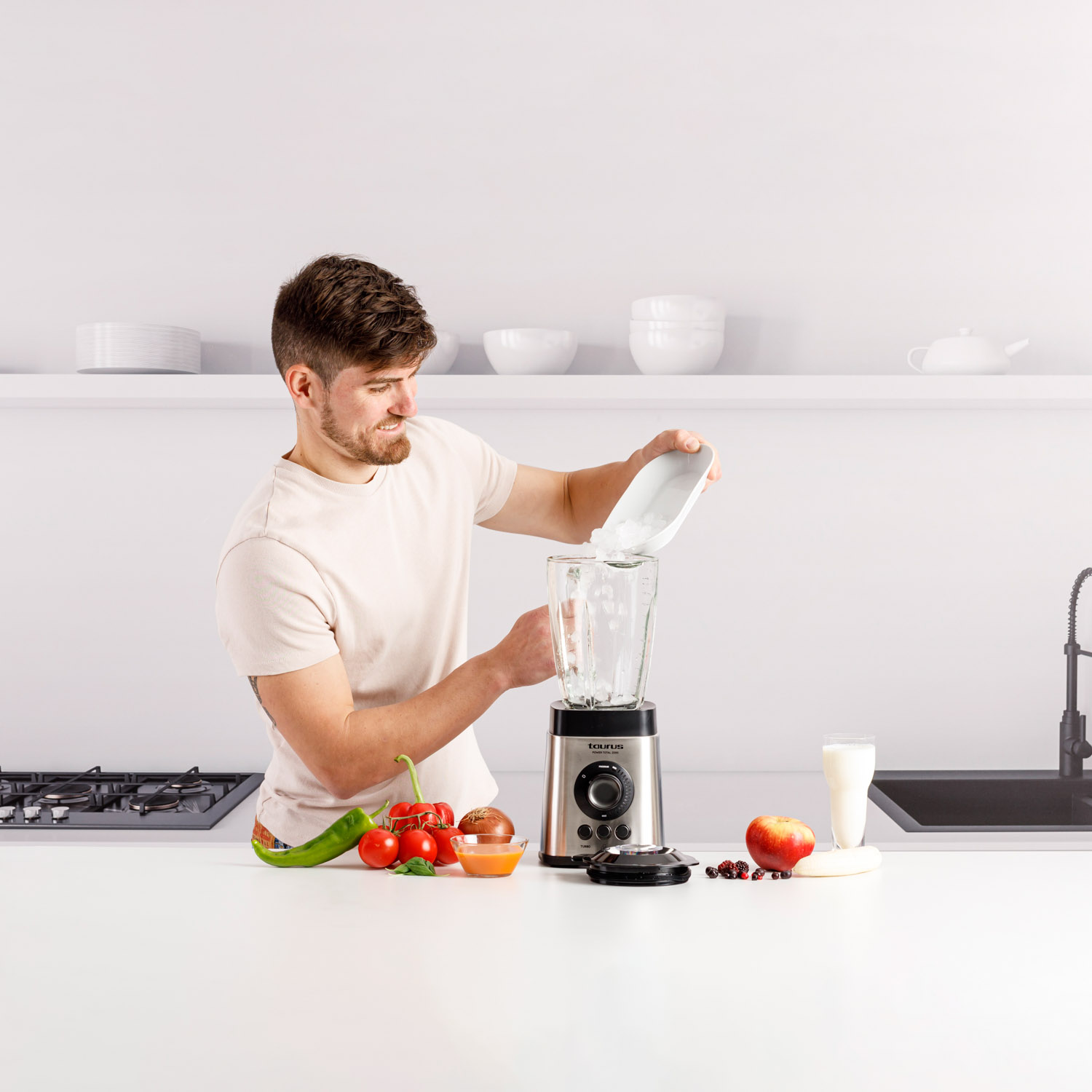 Un hombre vertiendo ingredientes en una licuadora, con verduras y frutas en la encimera.