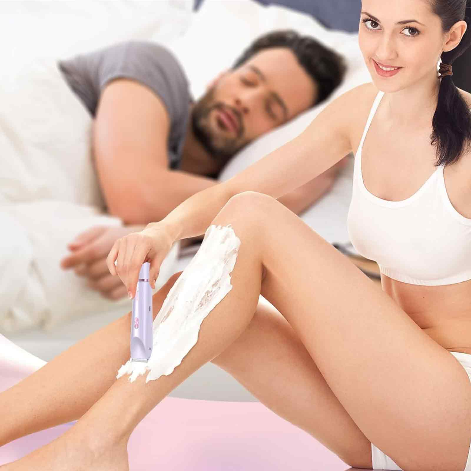 Frau rasiert Bein mit Epilierer, während eine andere Person hinter ihr im Bett schläft.
