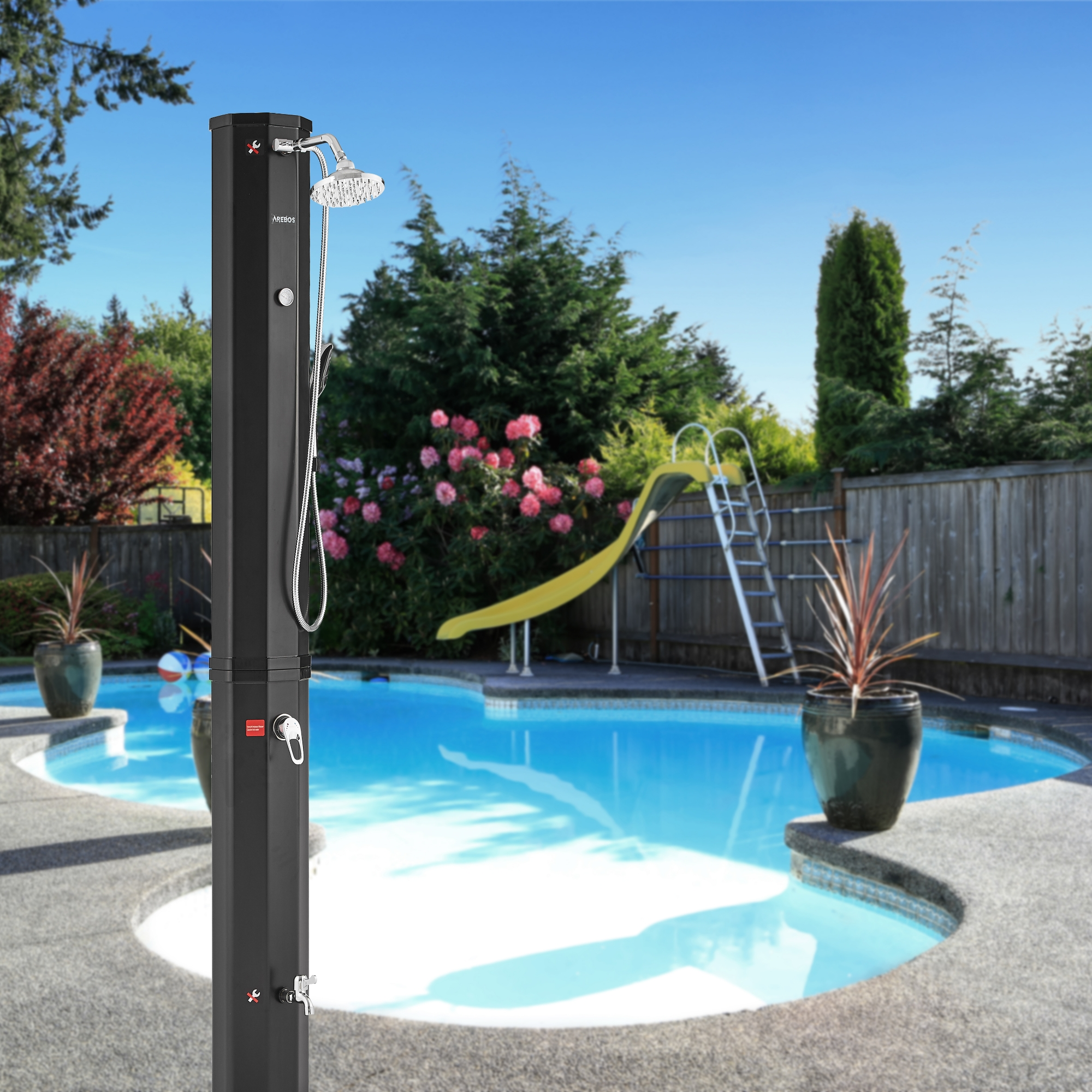 Schwarze Außendusche neben einem Pool mit Rutsche und Bäumen im Hintergrund, unter blauem Himmel.