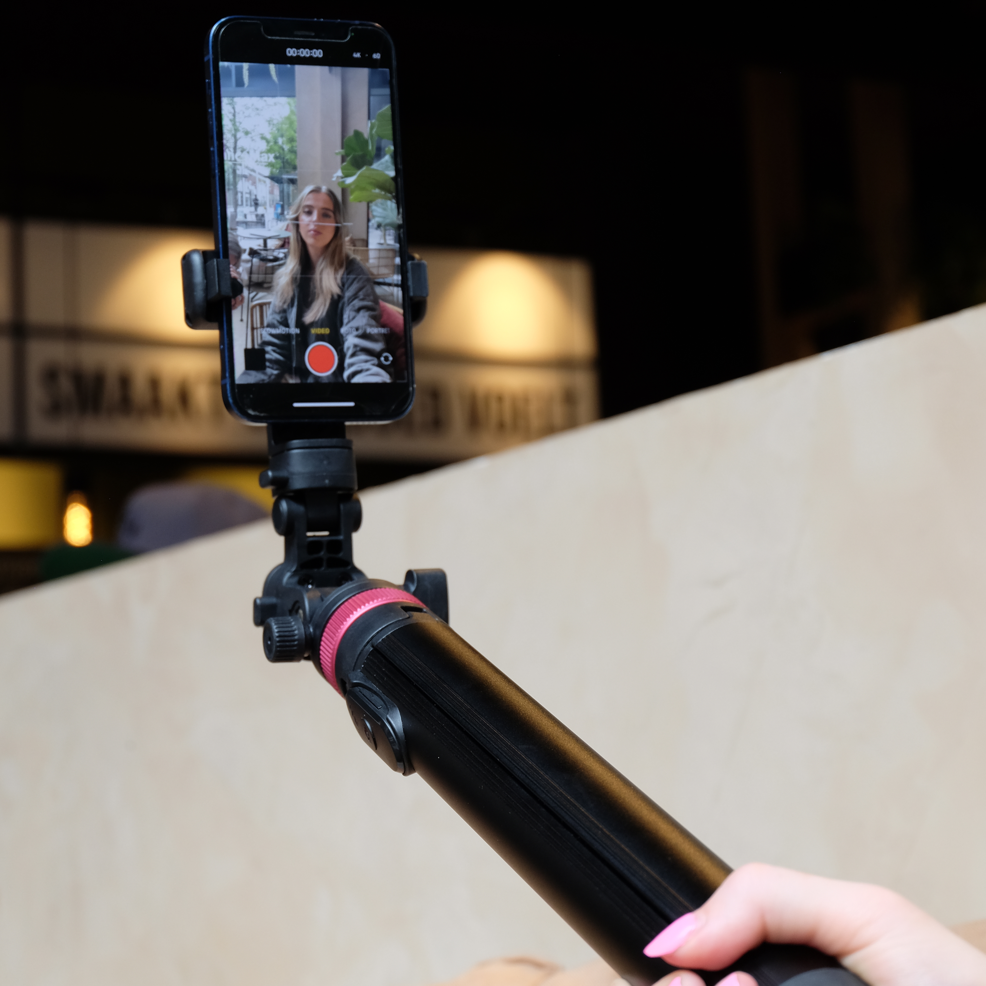 Une personne tient un téléphone sur une perche à selfie, enregistrant une vidéo. Le téléphone affiche une femme. Le fond est un café.