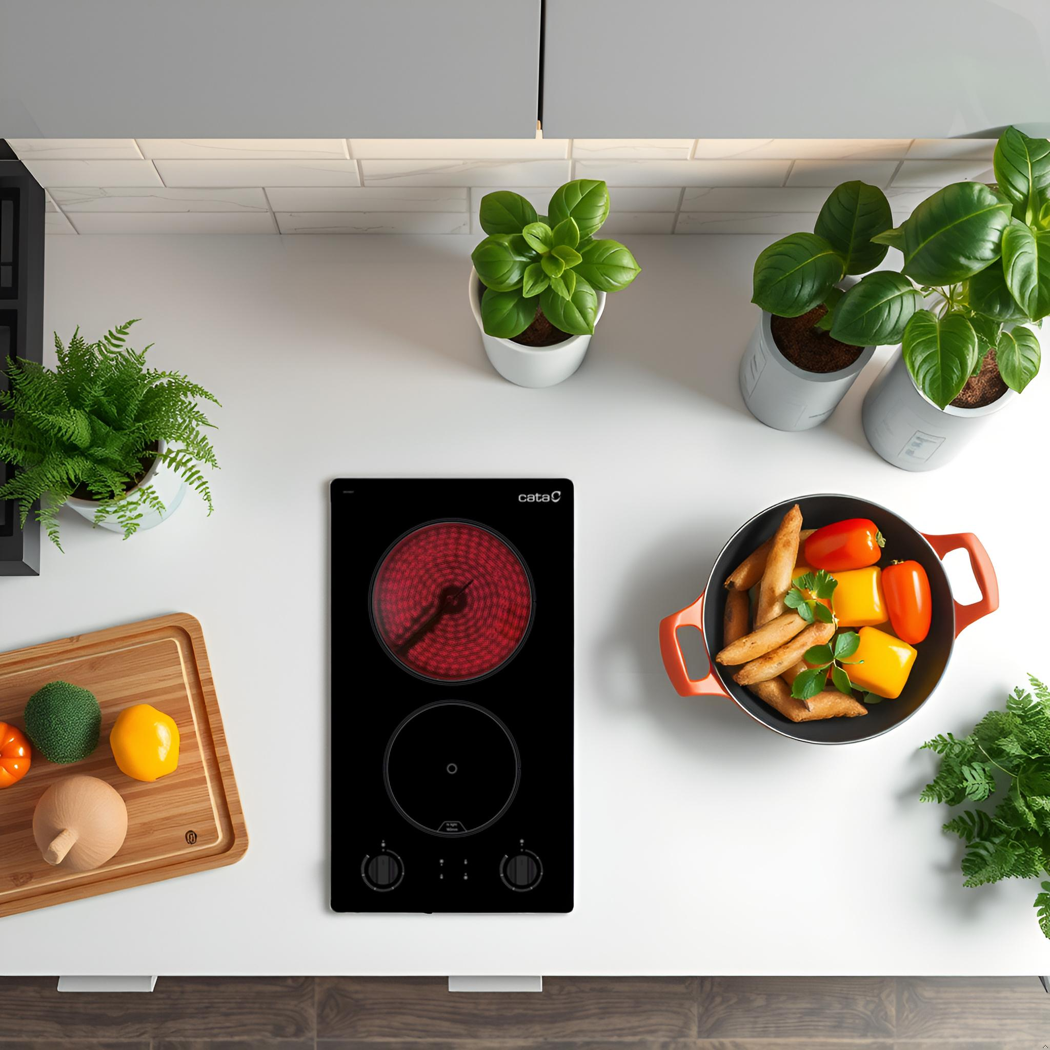 Vista superior: placa de inducción, verduras, plantas y tabla de cortar sobre una encimera blanca.