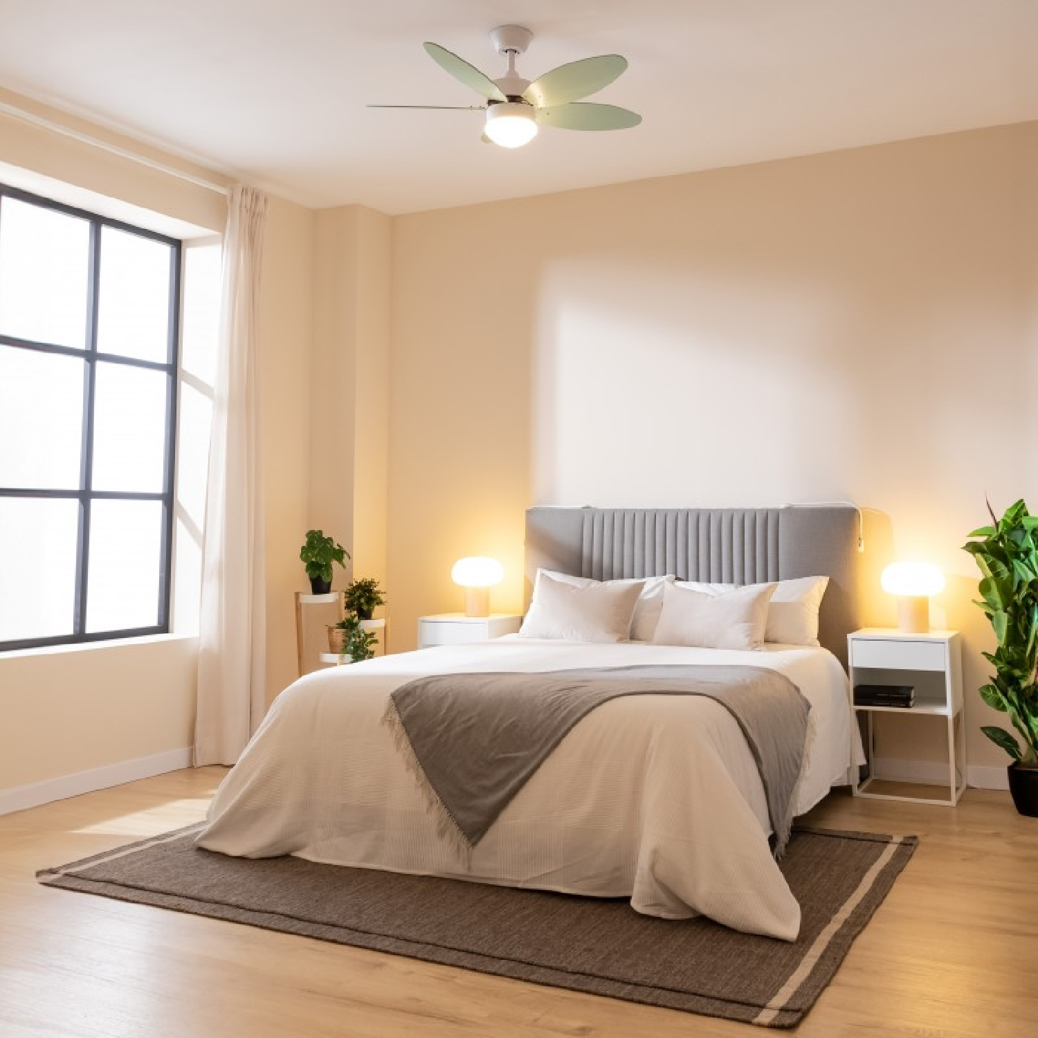 Sypialnia z łóżkiem, oknem, roślinami i wentylatorem sufitowym. Ściany są beżowe. Łóżko ma białą pościel z szarym narzutem.