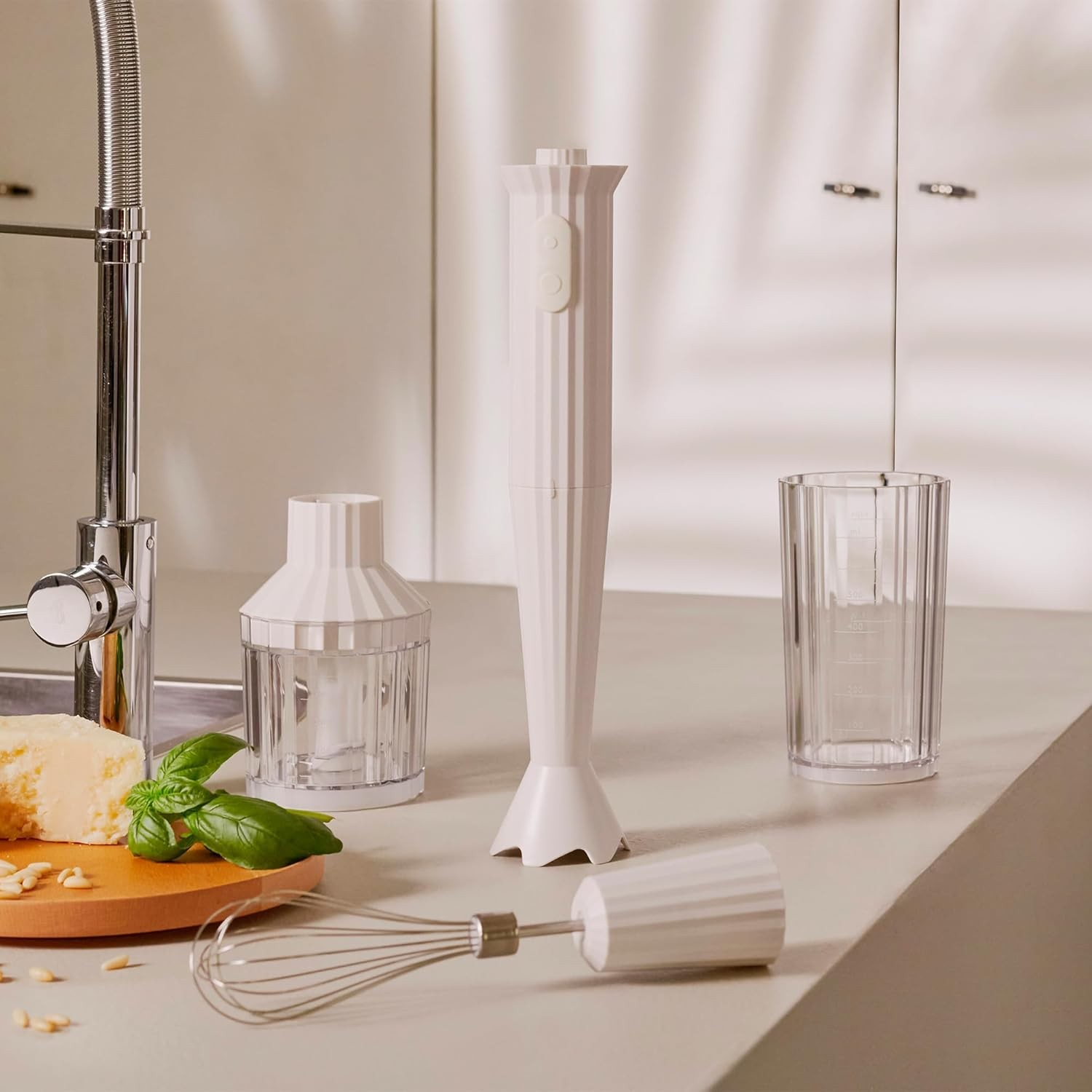 Frullatore a immersione bianco con accessori e ingredienti su un piano cucina. Visibile un lavandino.