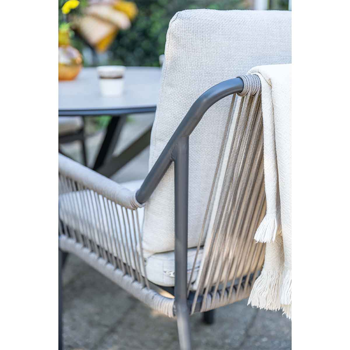 A chair with a white blanket. The chair is gray and made of a woven material. A table and a cup are in the background.