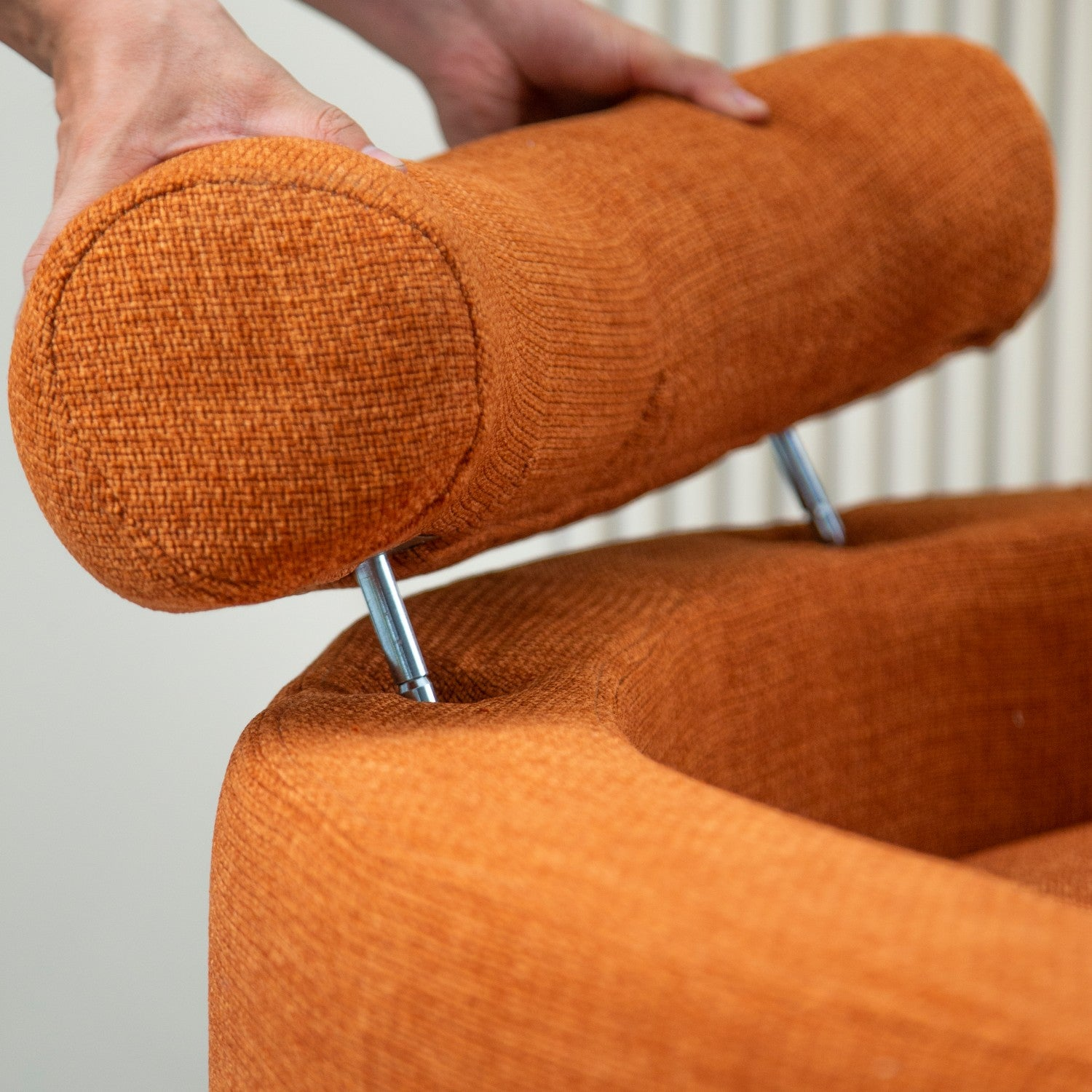 Manos sosteniendo un reposacabezas naranja. El reposacabezas es cilíndrico y está en una silla.