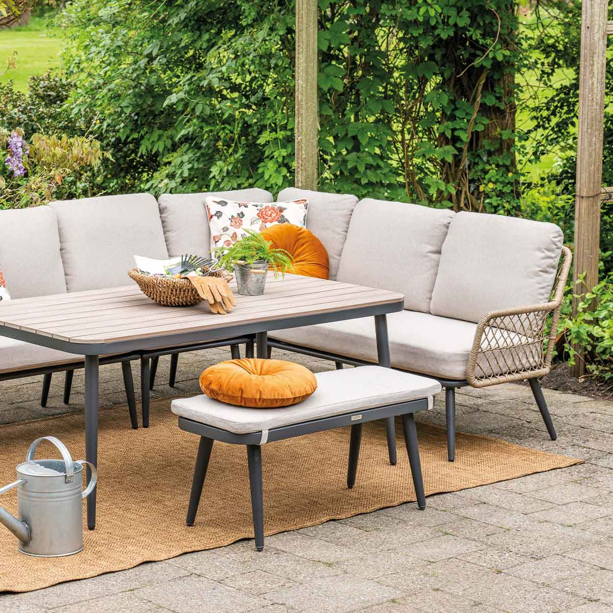 Outdoor patio set: table, bench, sofa with cushions, and watering can. Green background.