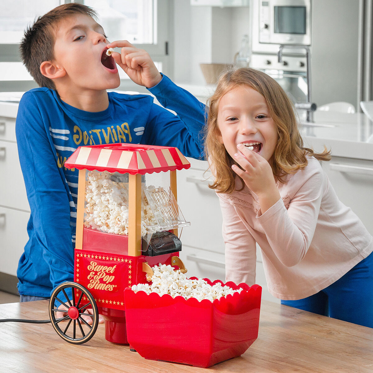 Zwei lächelnde Kinder essen Popcorn neben einer Popcornmaschine auf einem Holztisch.
