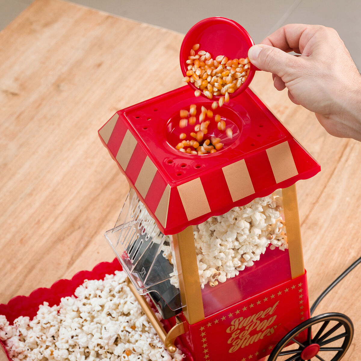 Hand gießt Maiskörner in rote Popcornmaschine mit gestreiftem Baldachin.