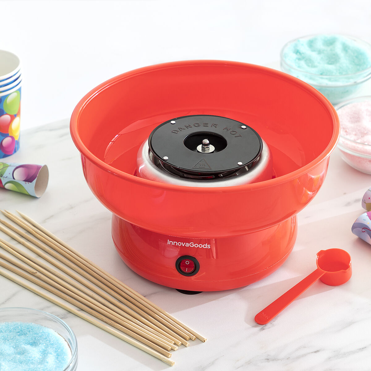 Eine rote Zuckerwattemaschine, ein Löffel und Stöcke. Schalen mit Zucker sind hinten.