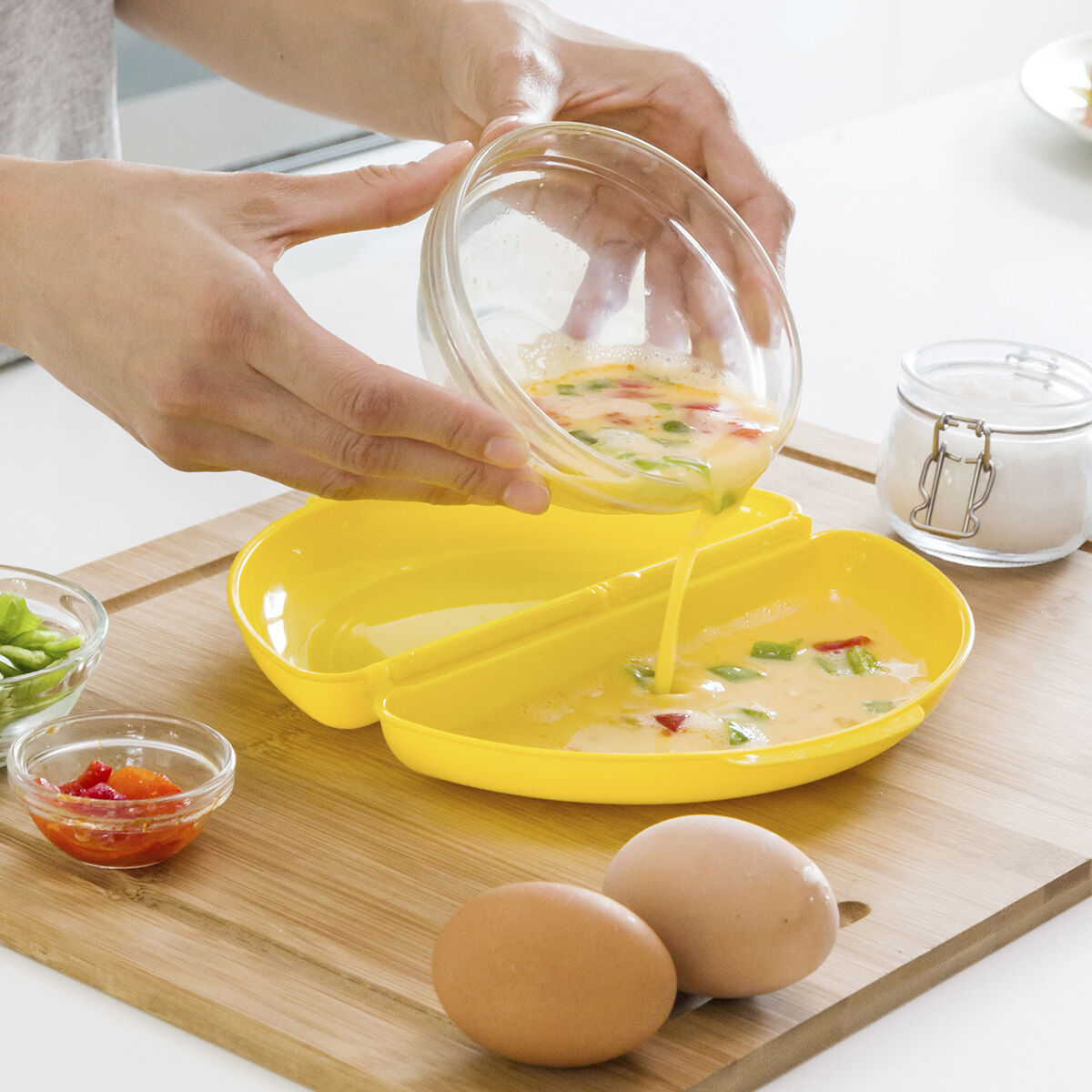 Manos vertiendo mezcla de huevo en una sartén amarilla de tortilla, con ingredientes.