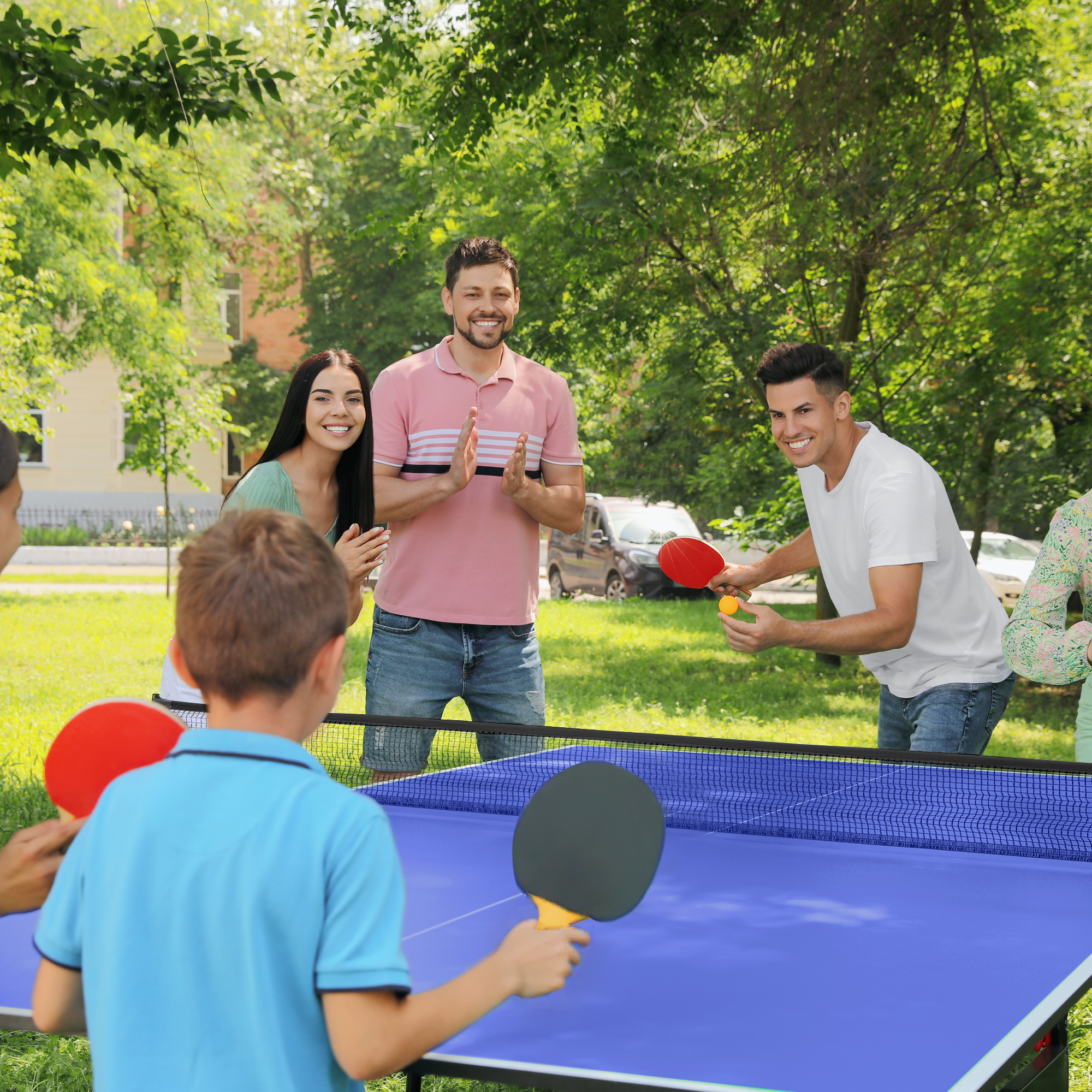 Gente jugando al tenis de mesa al aire libre. Un niño golpea la pelota. Otros miran y animan.