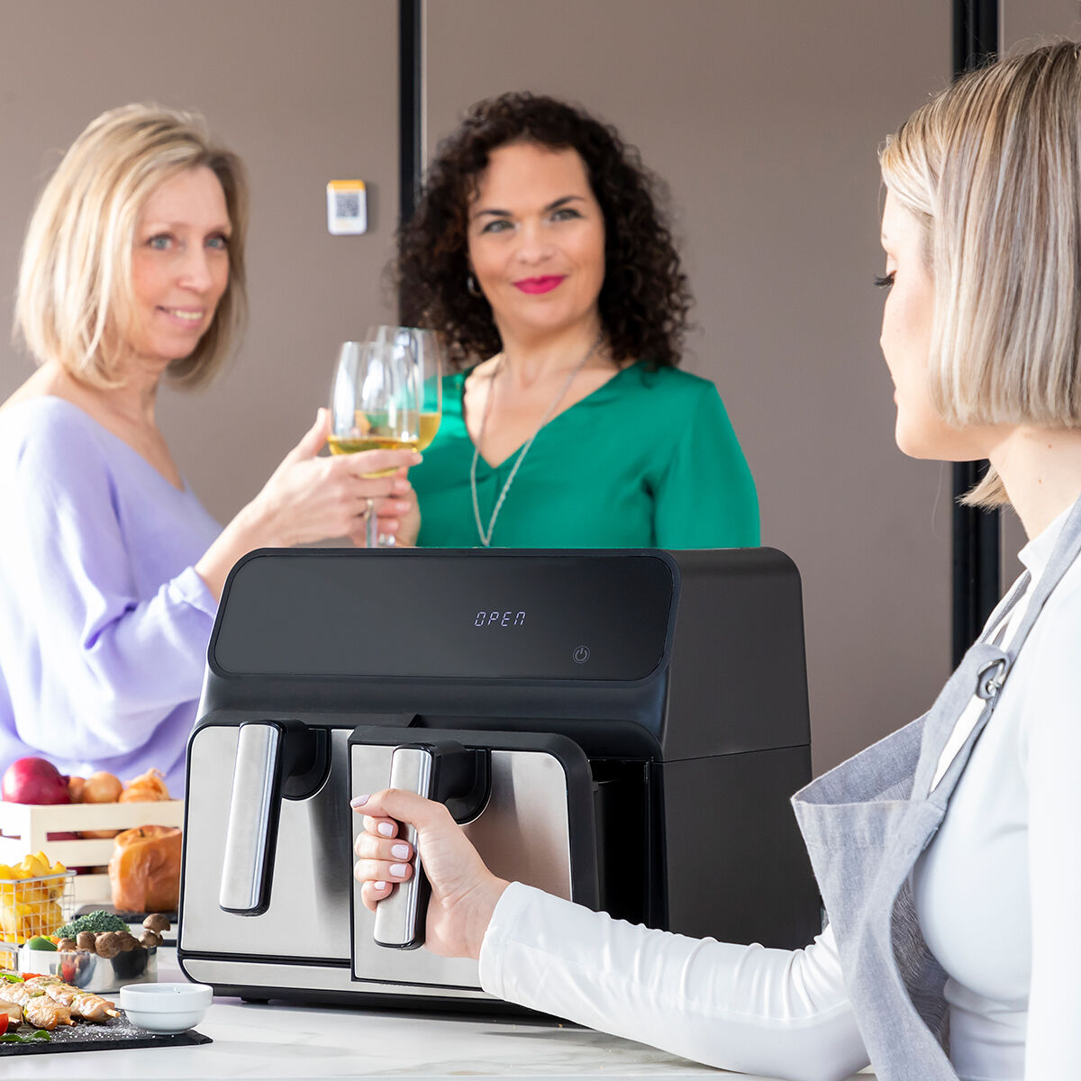 Drei Frauen stoßen mit Wein auf, in der Nähe einer Heißluftfritteuse mit Essen.