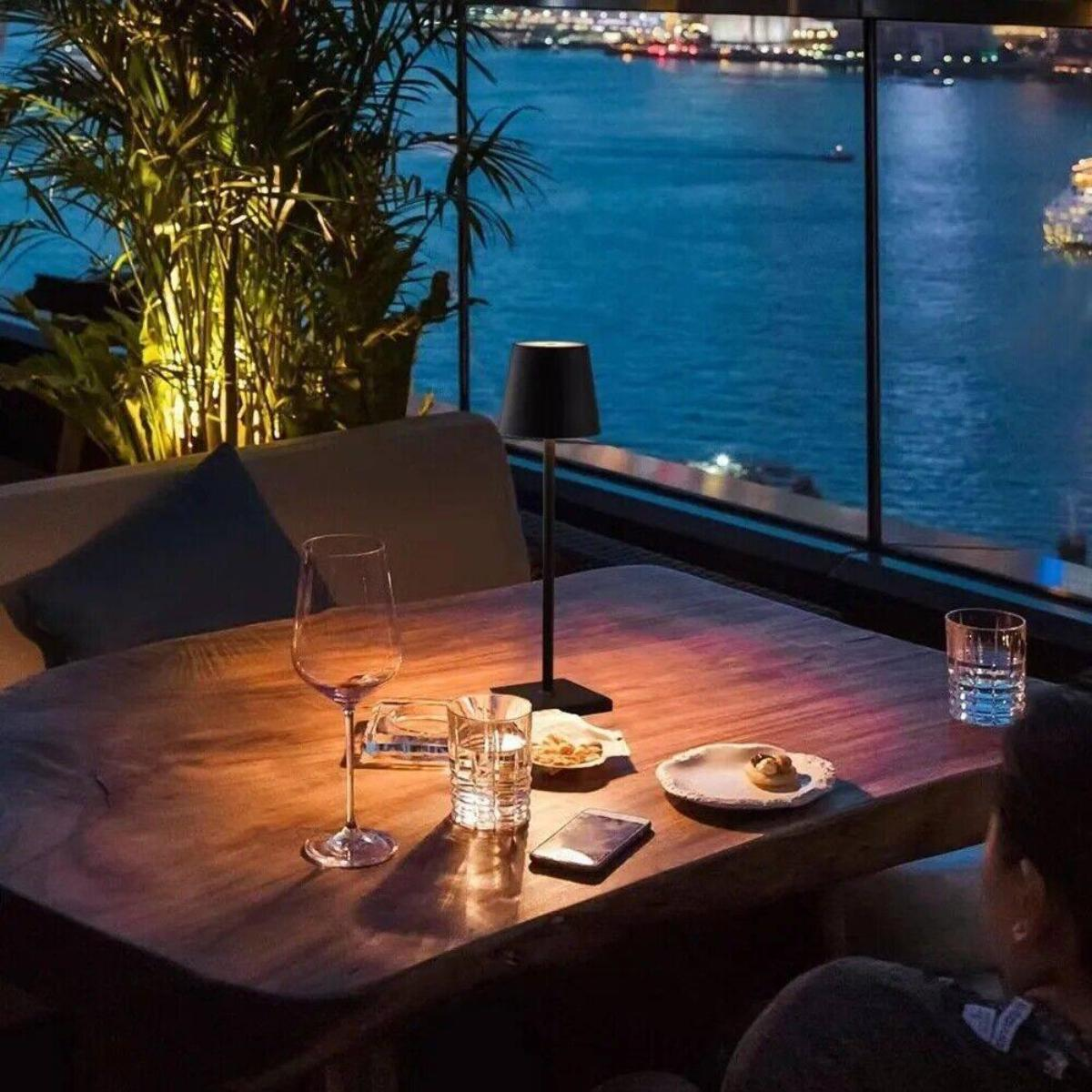 Ein Tisch mit Lampe, Weinglas und Snacks mit Blick auf eine Nachtstadt und das blaue Meer.