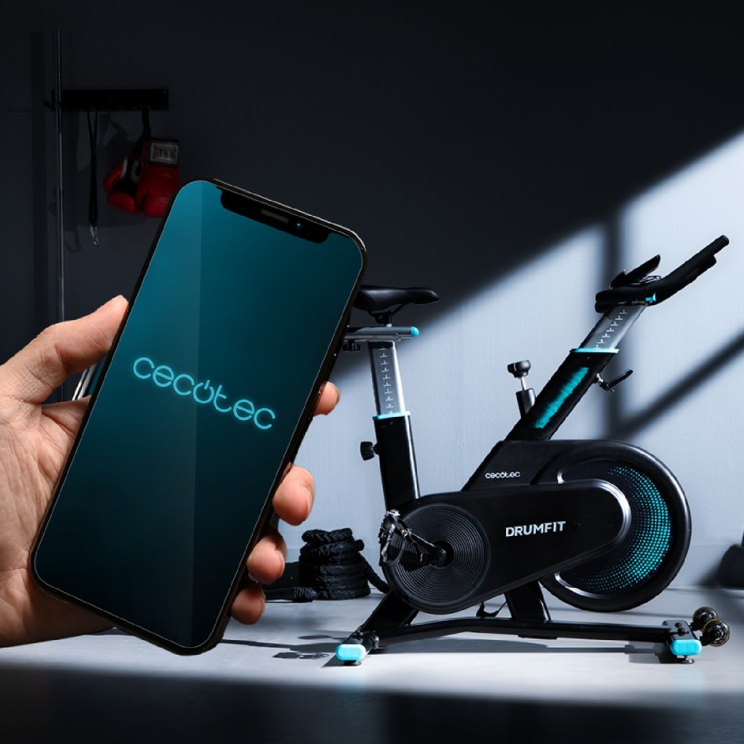 Una mano tiene uno smartphone con 'Cecotec' sullo schermo, vicino a una cyclette DRUMFIT.