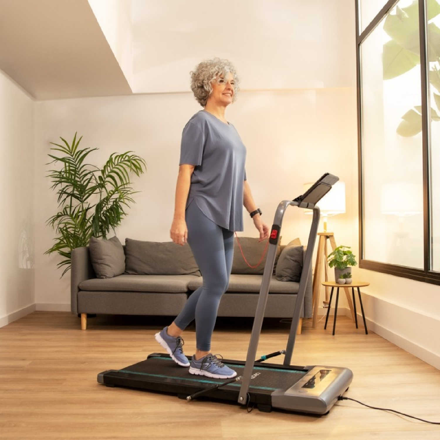 Starsza kobieta chodzi na bieżni. Ma na sobie niebieski strój do ćwiczeń. Okno i roślina w tle.