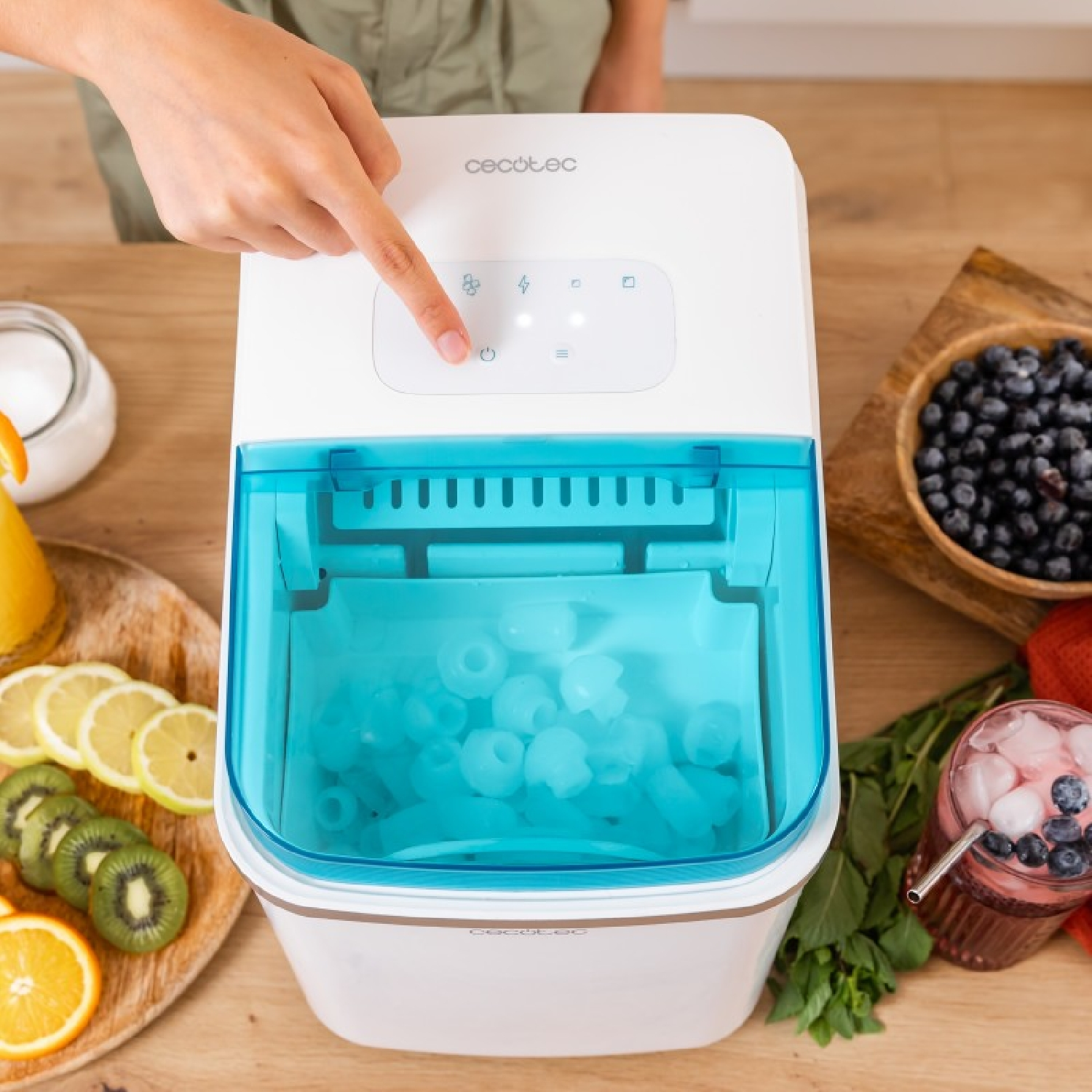 Eine Person drückt eine Taste auf einem weißen Eisbereiter. Eiswürfel und Obst sind in der Szene. Der Hintergrund ist Holz.