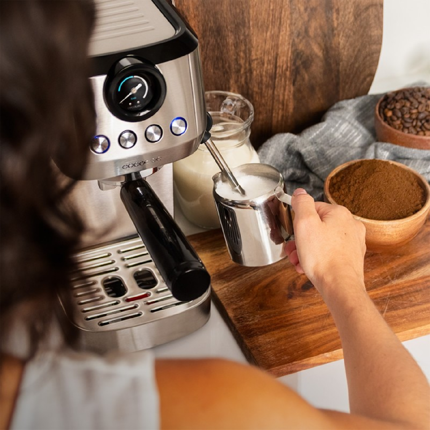 Osoba spienia mleko w stalowym dzbanku, ekspres do kawy i drewniana deska.