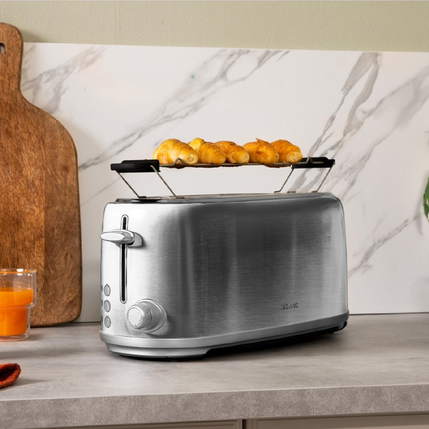 Srebrny toster z rogalikami na górze. Drewniana deska i szklanka soku w tle.