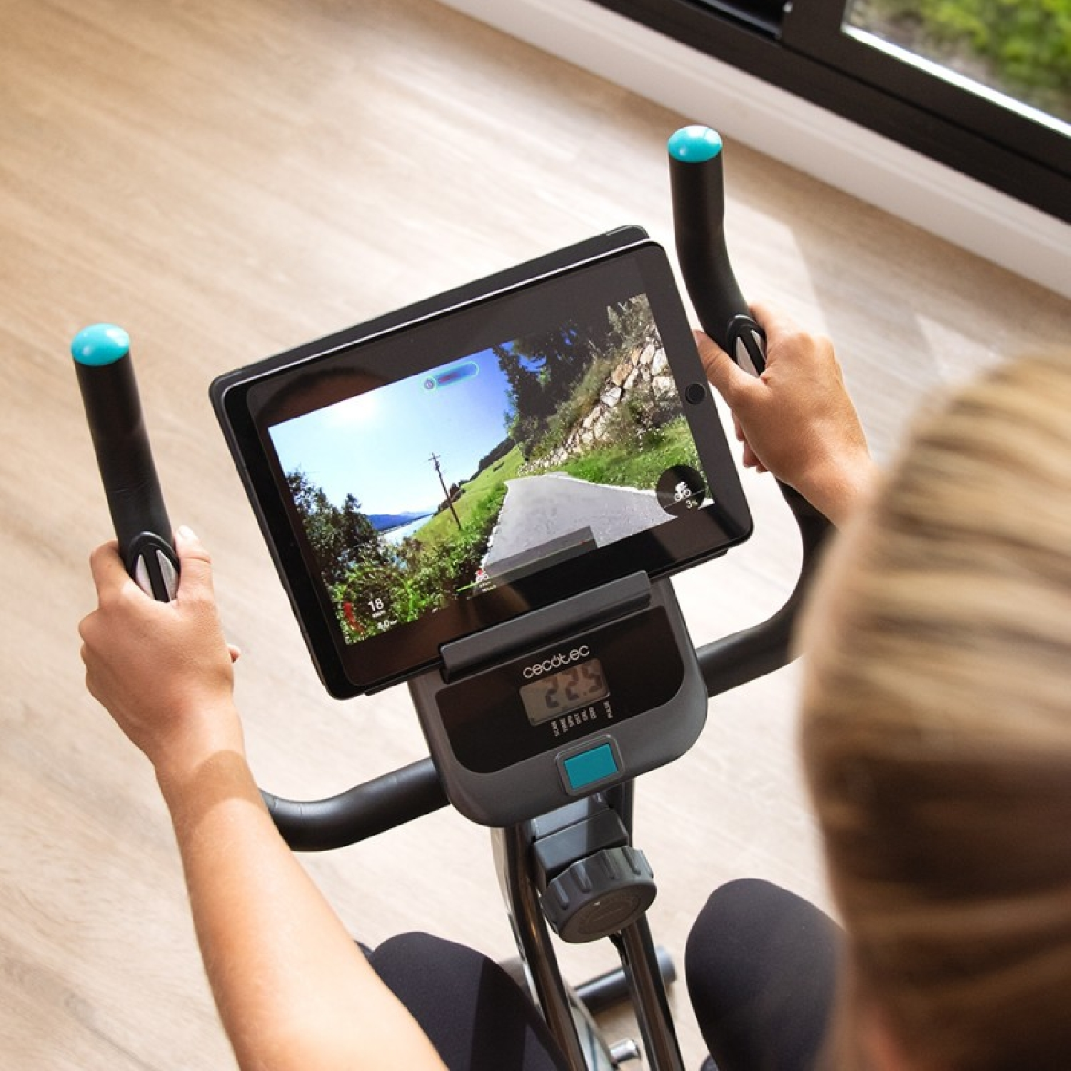 Kobieta korzystająca z roweru treningowego z tabletem wyświetlającym widok drogi. Rower ma niebieskie akcenty.