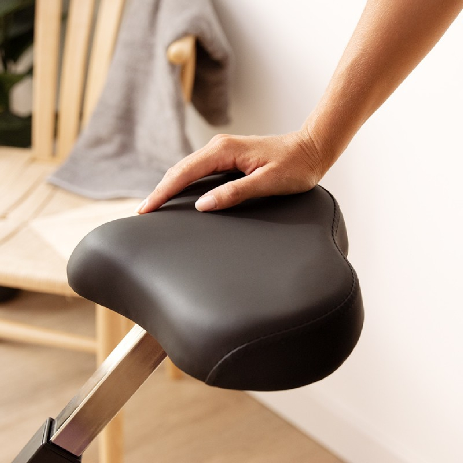 Dłoń spoczywa na czarnym siodełku rowerowym. W tle widoczna drewniana ławka i ręcznik.