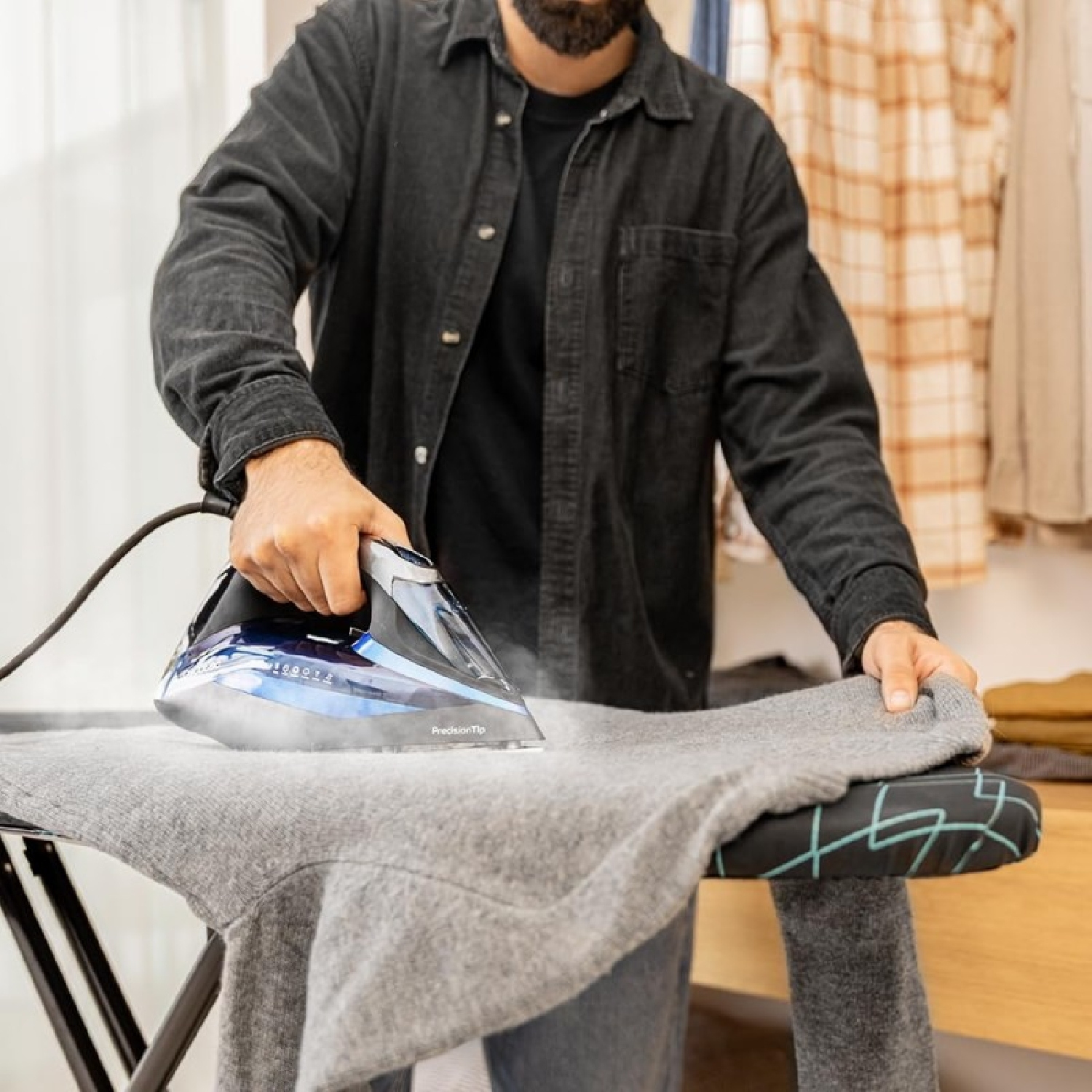 Uomo che stira un maglione grigio. Ferro nero con 'PrecisionTip'. Camicia marrone sullo sfondo.