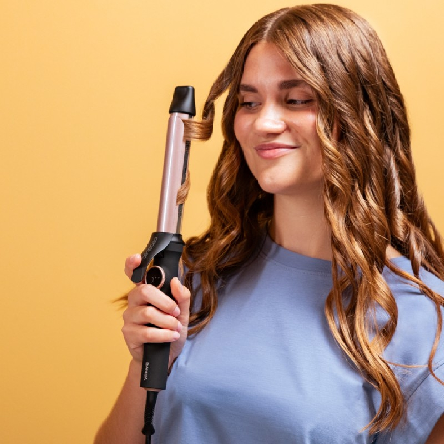 Mujer rizándose el pelo con un rizador. Sonríe, con un fondo amarillo.