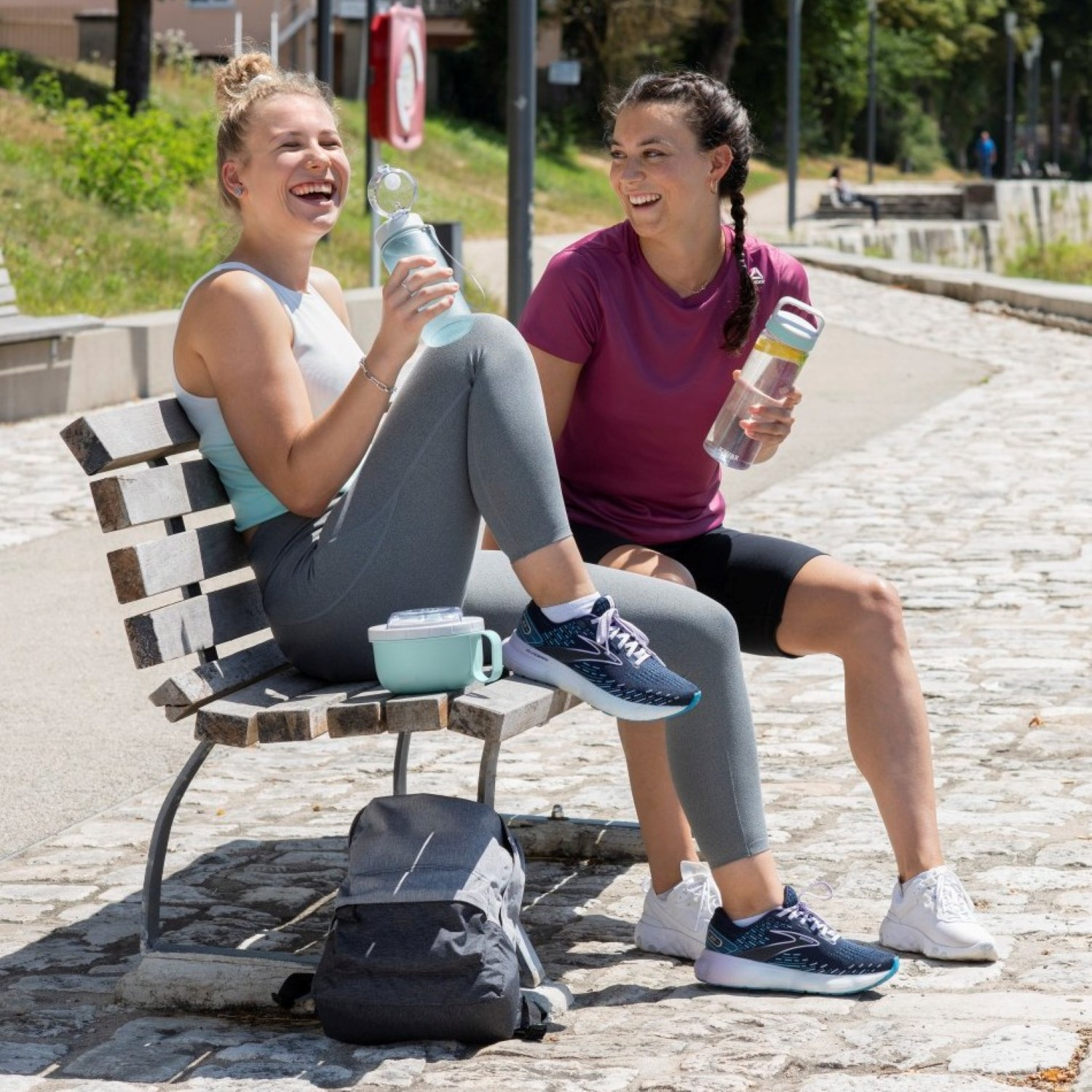 Zwei Frauen sitzen auf einer Bank, halten Wasserflaschen, lächeln und lachen. Graue Leggings und Turnschuhe.