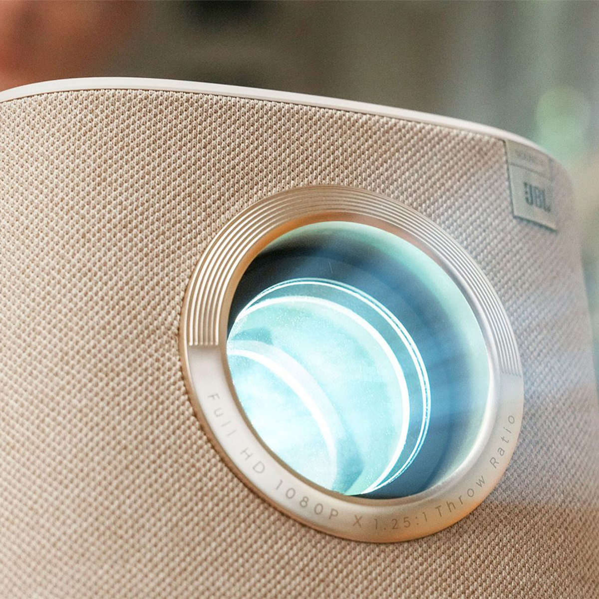 Projektor z okrągłym obiektywem jest podświetlony. Projektor ma beżowe wykończenie z tkaniny.